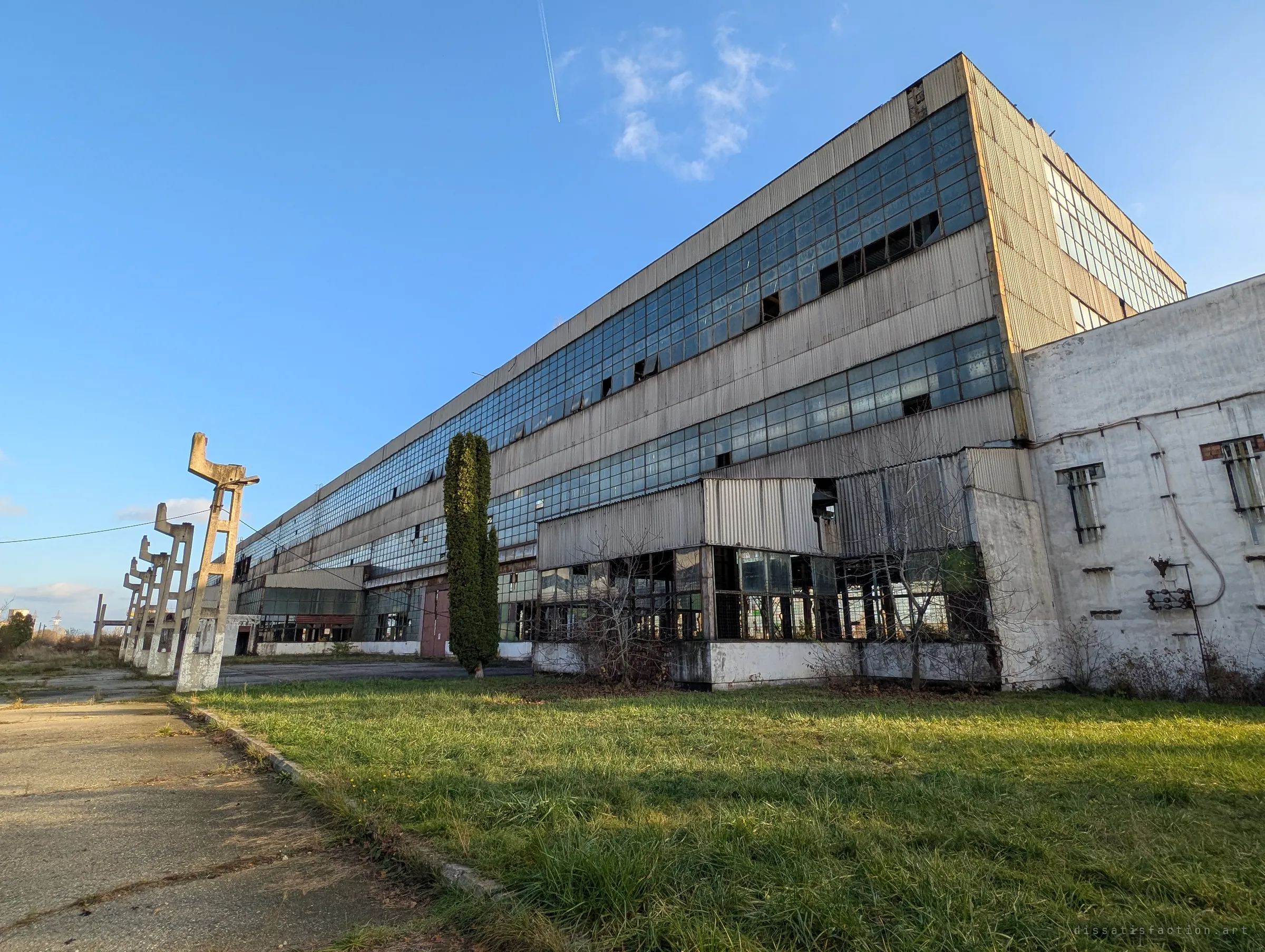 A vast industrial hall with long bands of broken glazing stretches away under a clear blue sky, a line of angular concrete lamp posts leaning along the path. The low wide angle shows the sheer scale of the complex.
