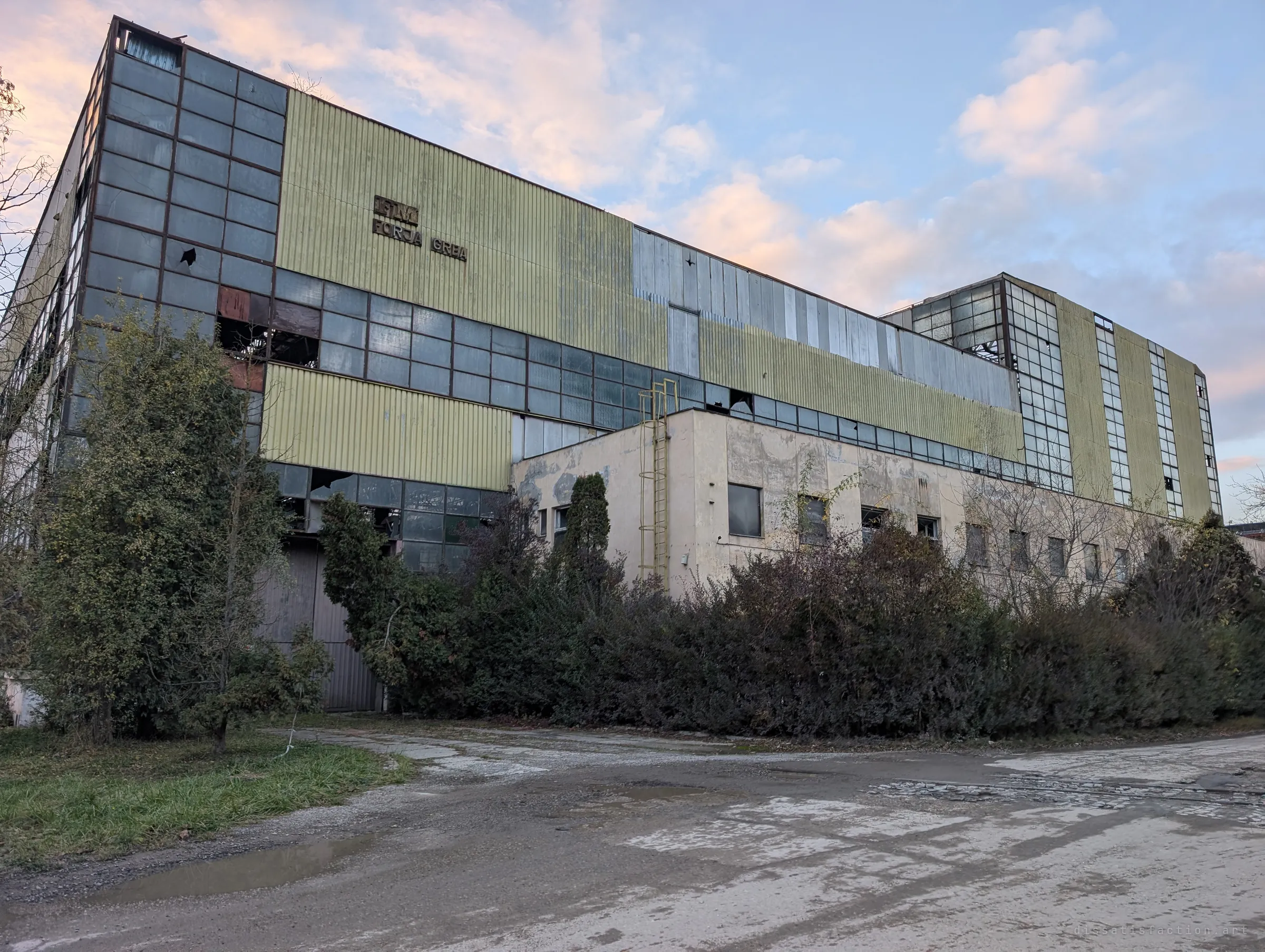A tall hall clad in faded green panels and grids of broken glass rises behind overgrown shrubs under a pink evening sky. The size of the building hints at the heavy work once done inside.