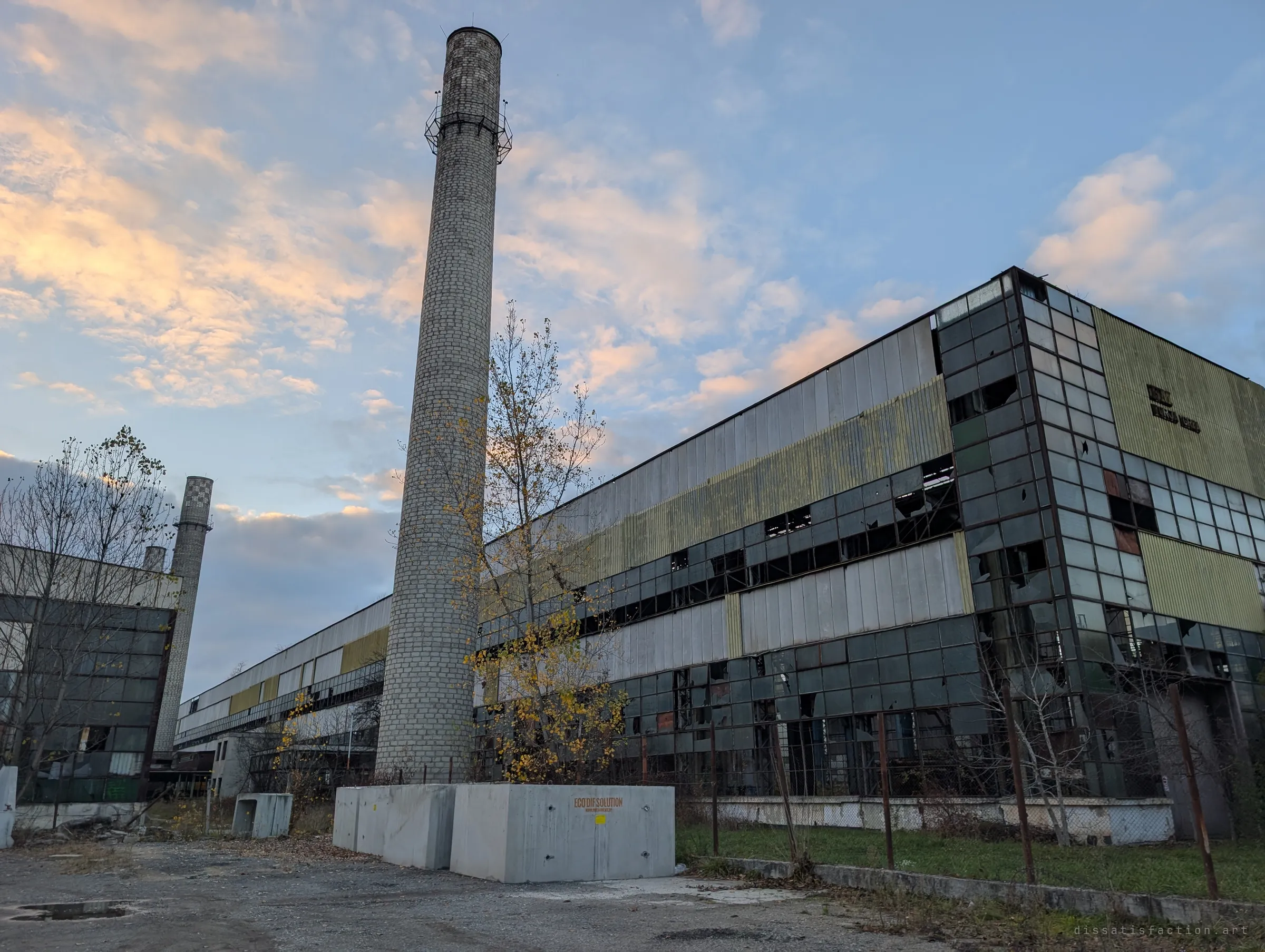 A tall brick chimney rises beside a long hall of broken glass and faded green cladding, lit by a pink dusk sky. The scale of the works is clear from the outside, before the doors.