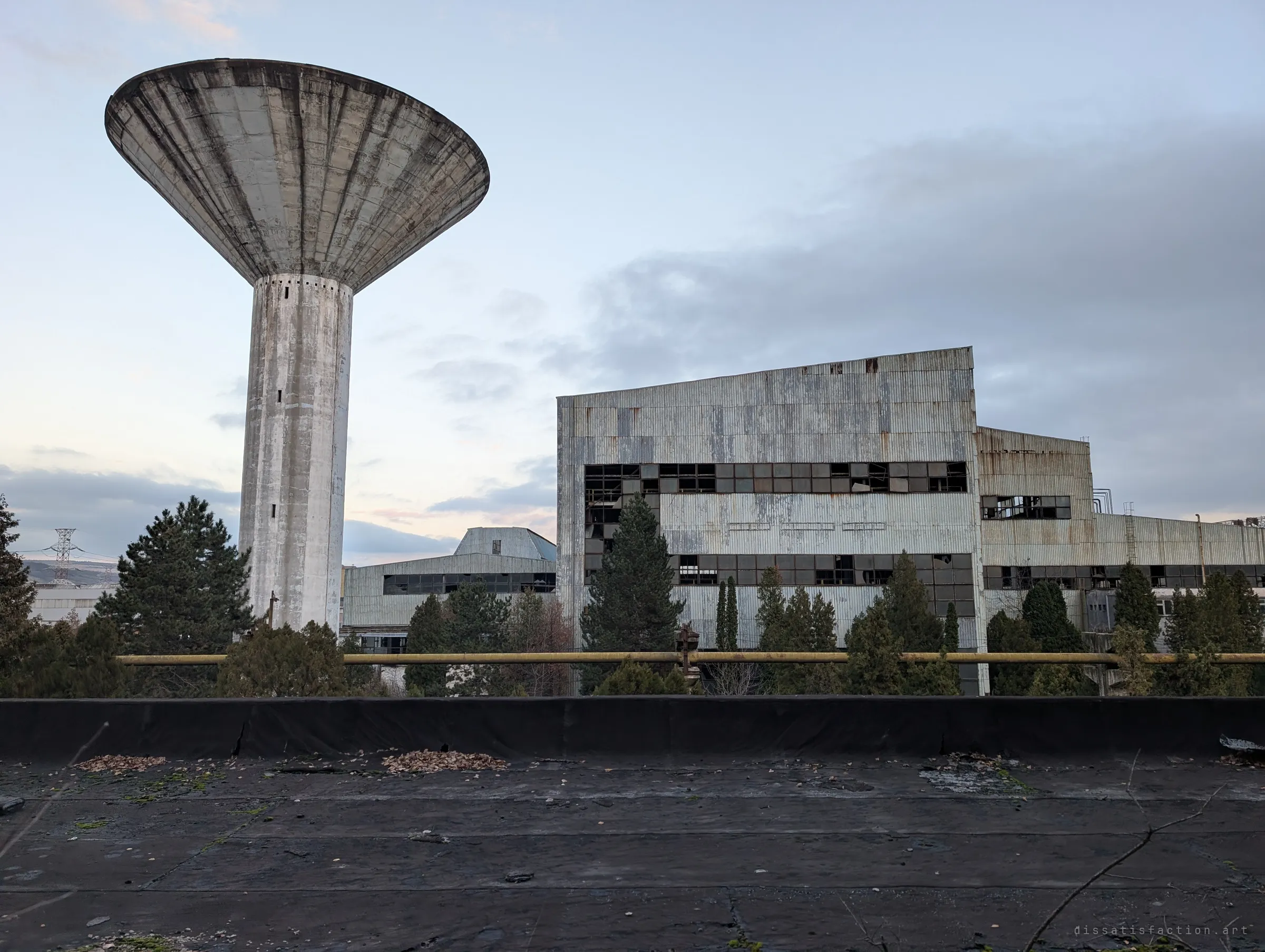 A mushroom-shaped concrete water tower stands over pine trees and a corrugated factory block, seen from a rooftop at dusk. It is the silhouette that marks the whole complex.