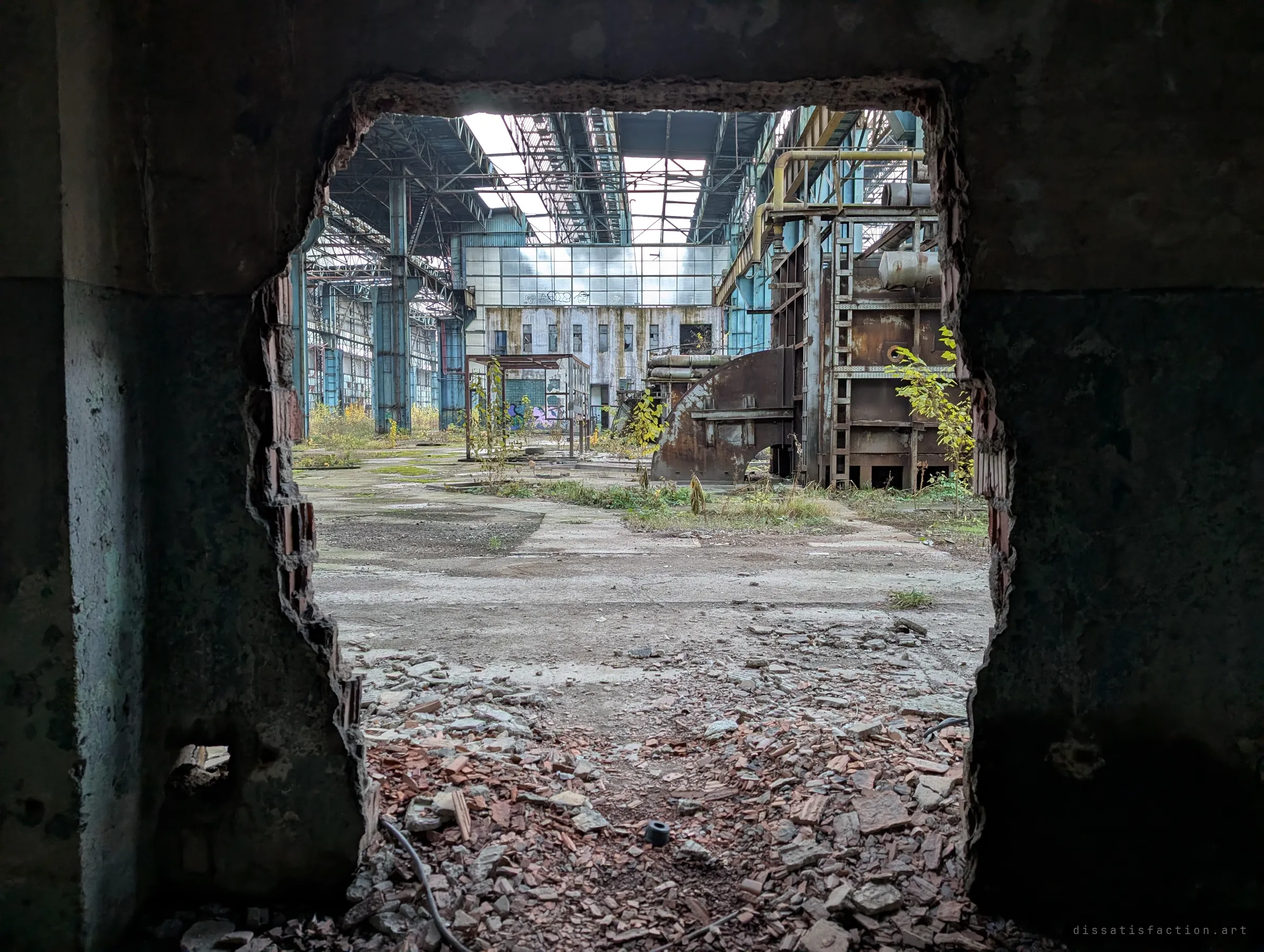A rough hole smashed through a concrete wall frames a vast, overgrown factory hall with rusted furnaces and a glass end wall. The broken opening is the way in.