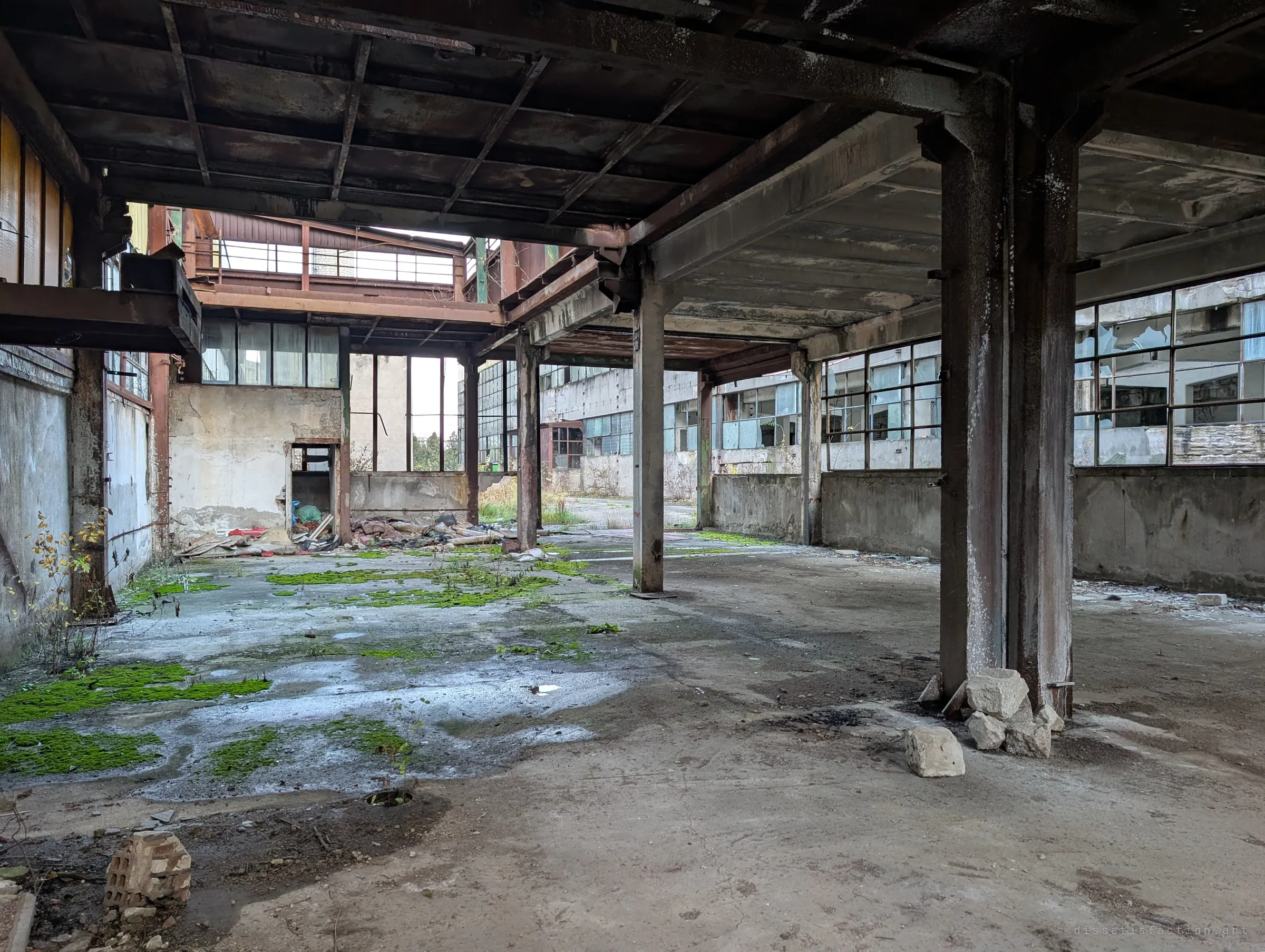 A low-ceilinged steel-framed bay with moss spreading across a wet concrete floor, broken windows on the right. Debris piles in the far corner under a mezzanine.
