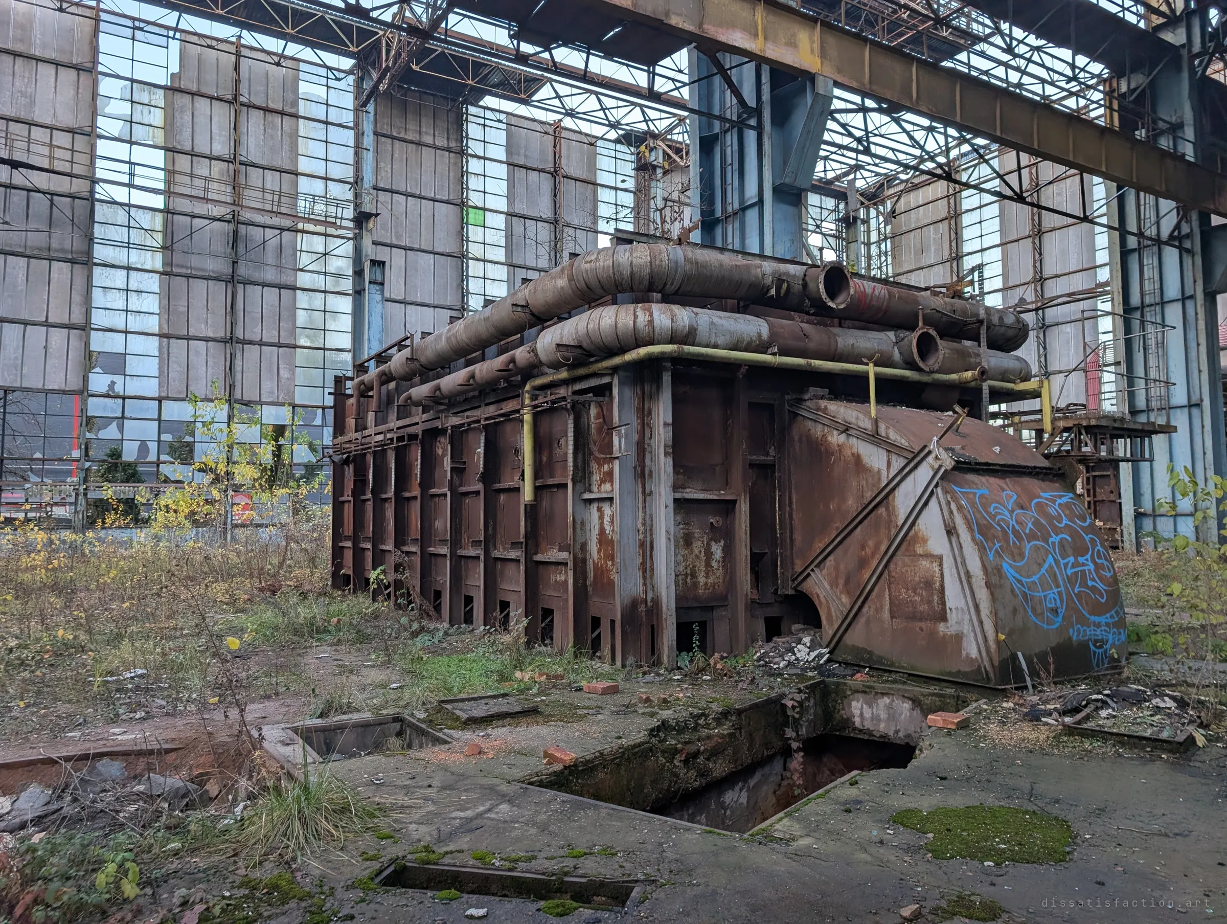 A huge rusted furnace wrapped in insulated ducts sits over open floor pits, with graffiti on its hood and a glass curtain wall behind. Weeds grow right up to the machine.