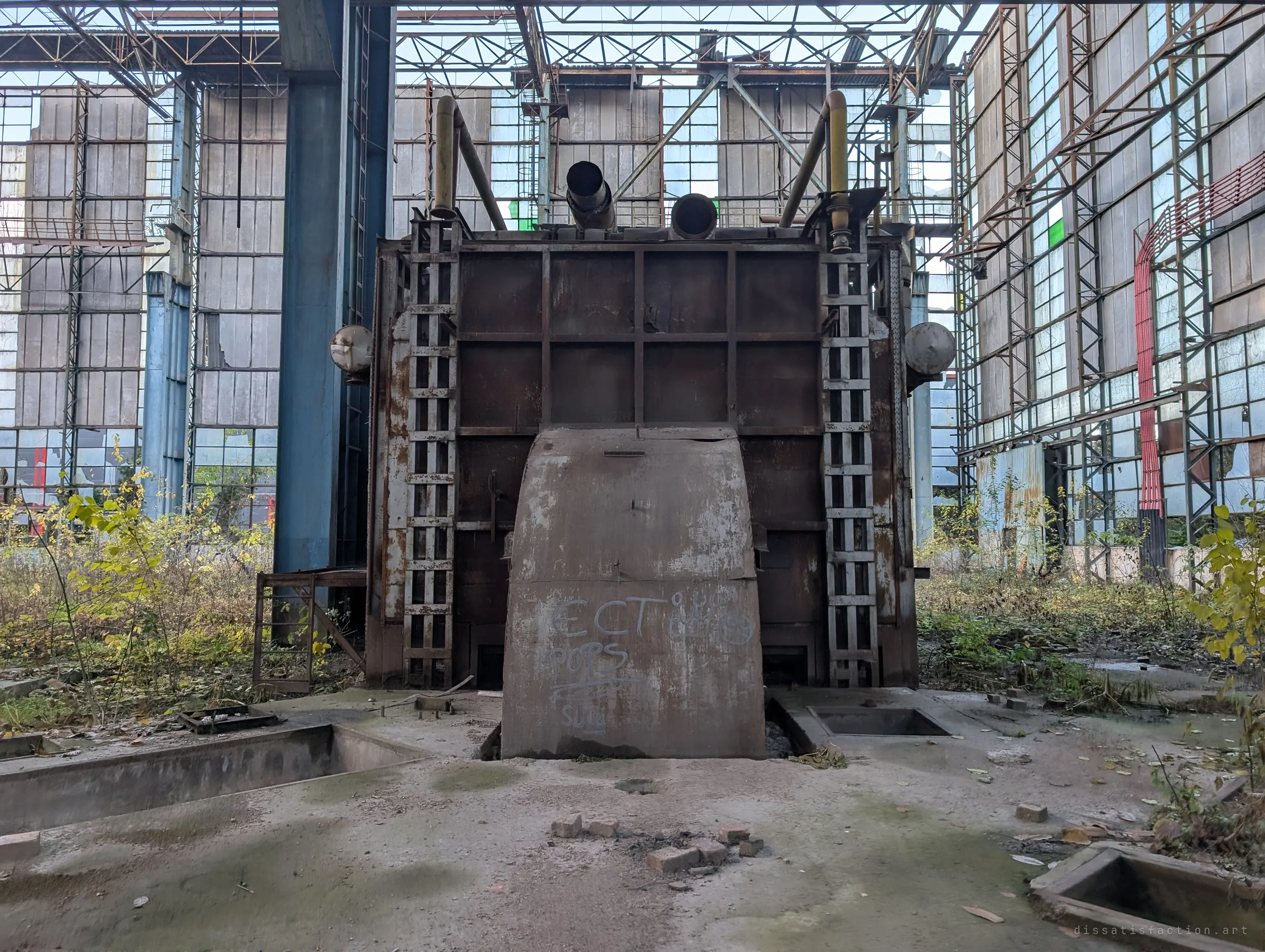 Head-on view of a rusted furnace with a curved steel hood and ladders on both sides, framed by the blue columns and glass walls of the hall. Brick scraps lie scattered in front.