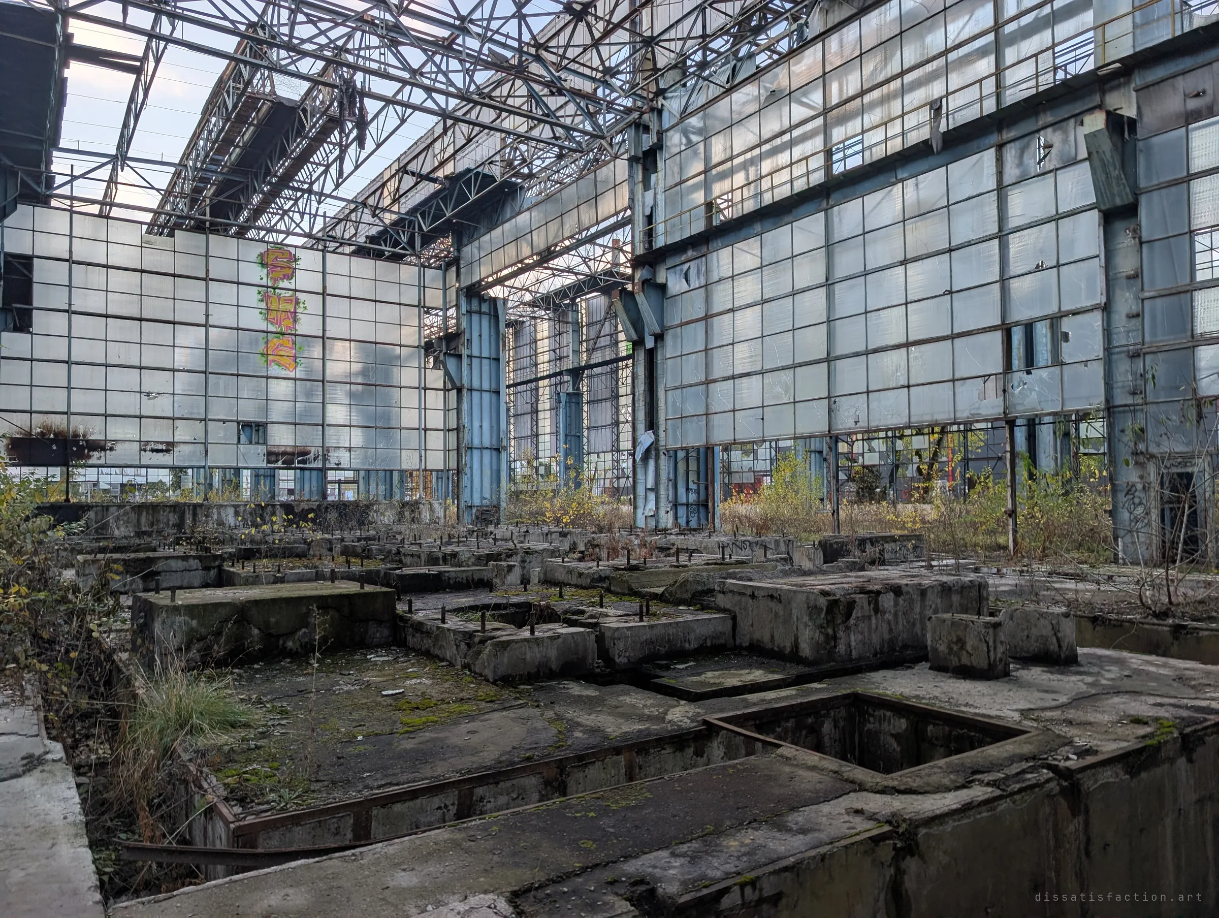 Rows of bare concrete machine foundations with bolts sticking out fill a hall of towering glass walls and open roof trusses. Colourful graffiti climbs one of the glass panels.