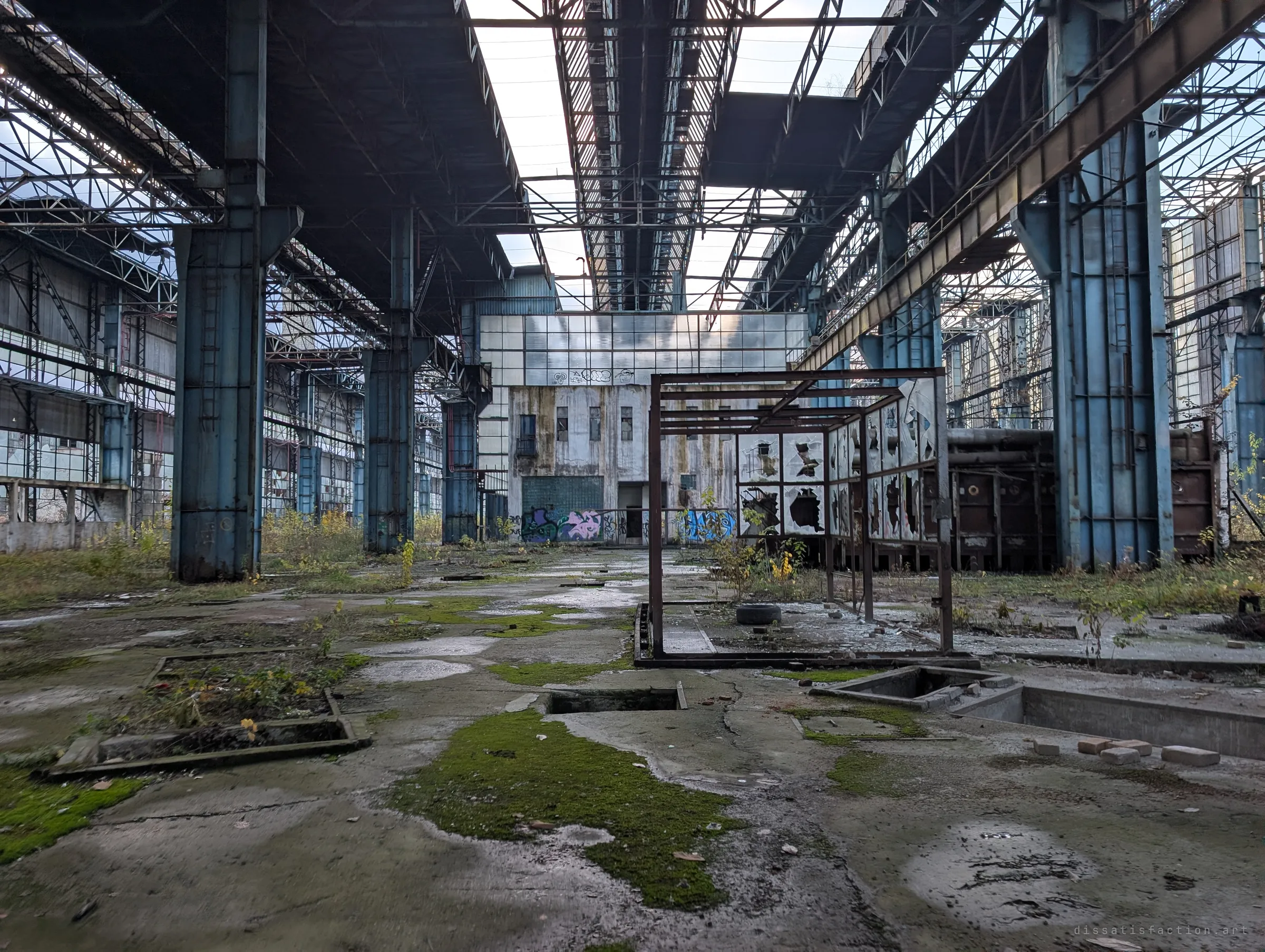 A wide, moss-patched hall floor leads between blue columns to a graffitied end building, with an empty steel frame and shattered panels in the middle. The open roof lets daylight pour in.