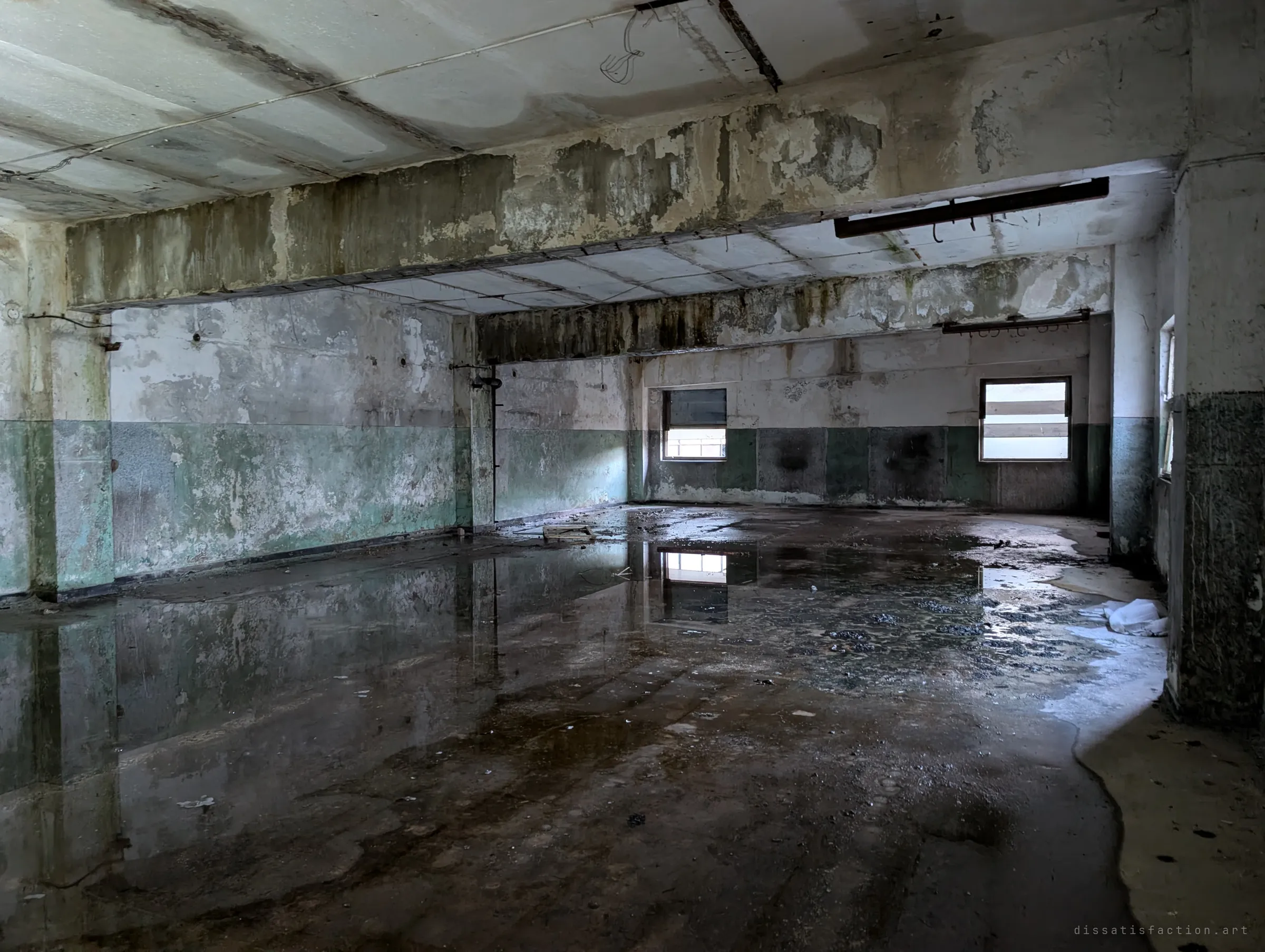 A flooded concrete room with peeling green and white walls, two small windows reflected in the standing water. Damp stains run across the ceiling beams.