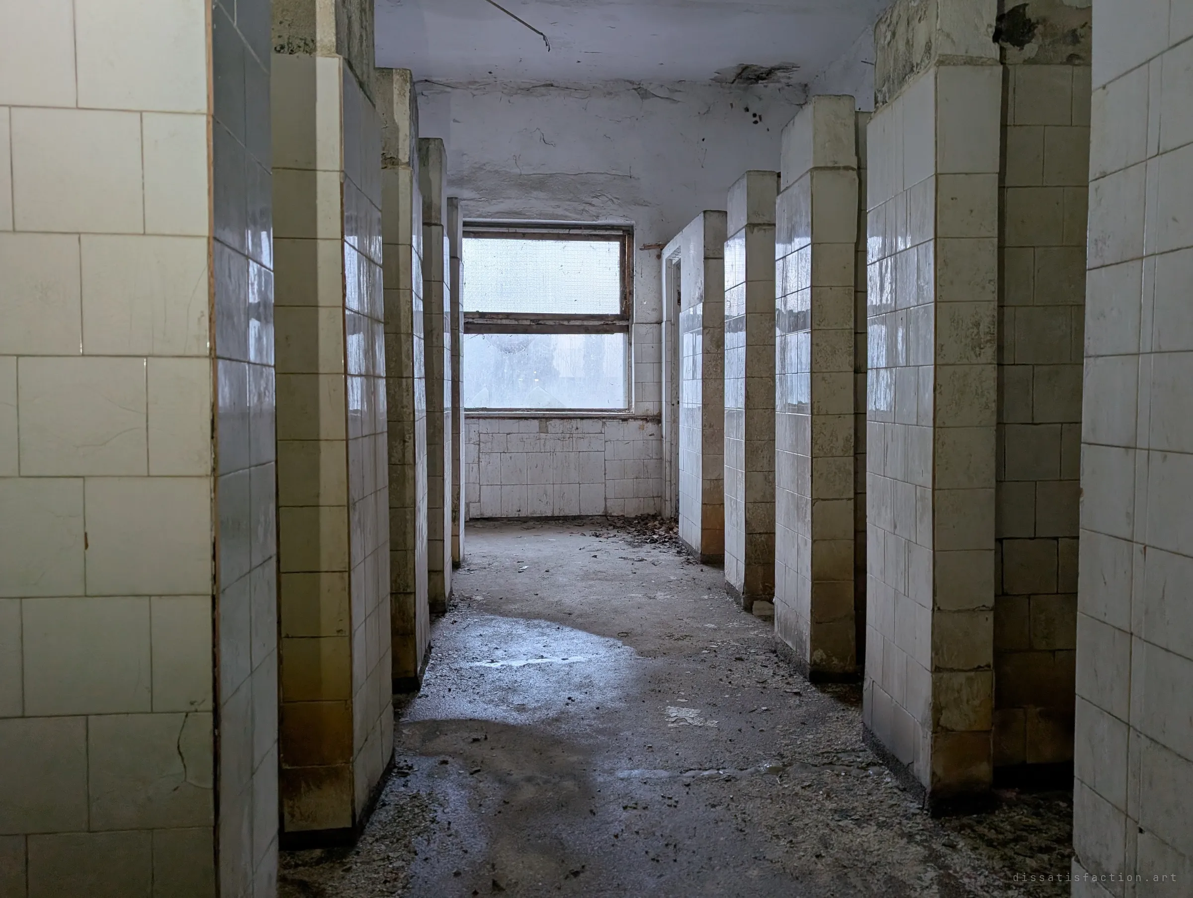 A row of tiled shower stalls lines a wet corridor toward a frosted window, the washroom of a workers’ amenity block left to decay.