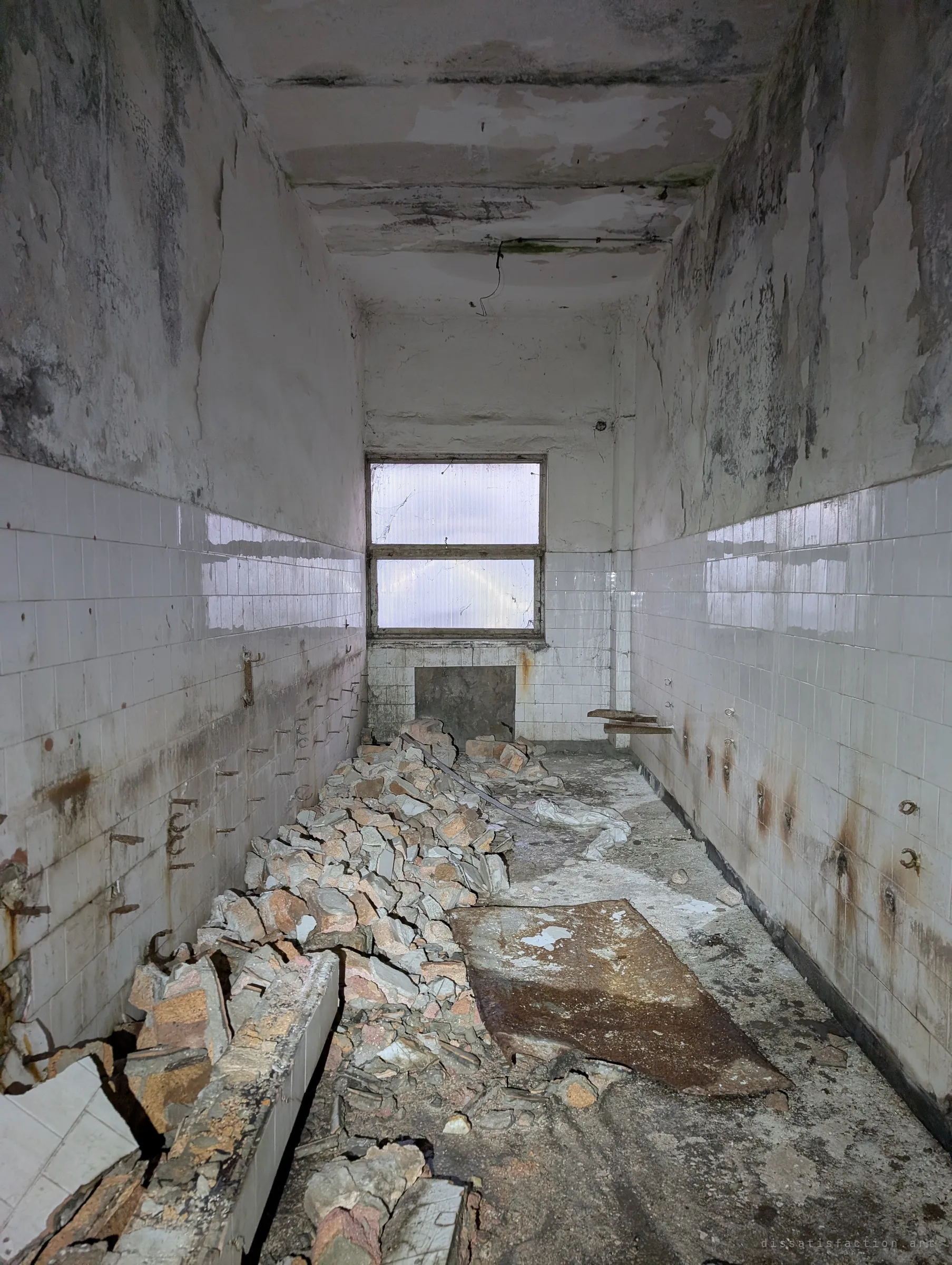 A narrow tiled washroom half filled with rubble, rusted pipe stubs running along both walls under a peeling ceiling.