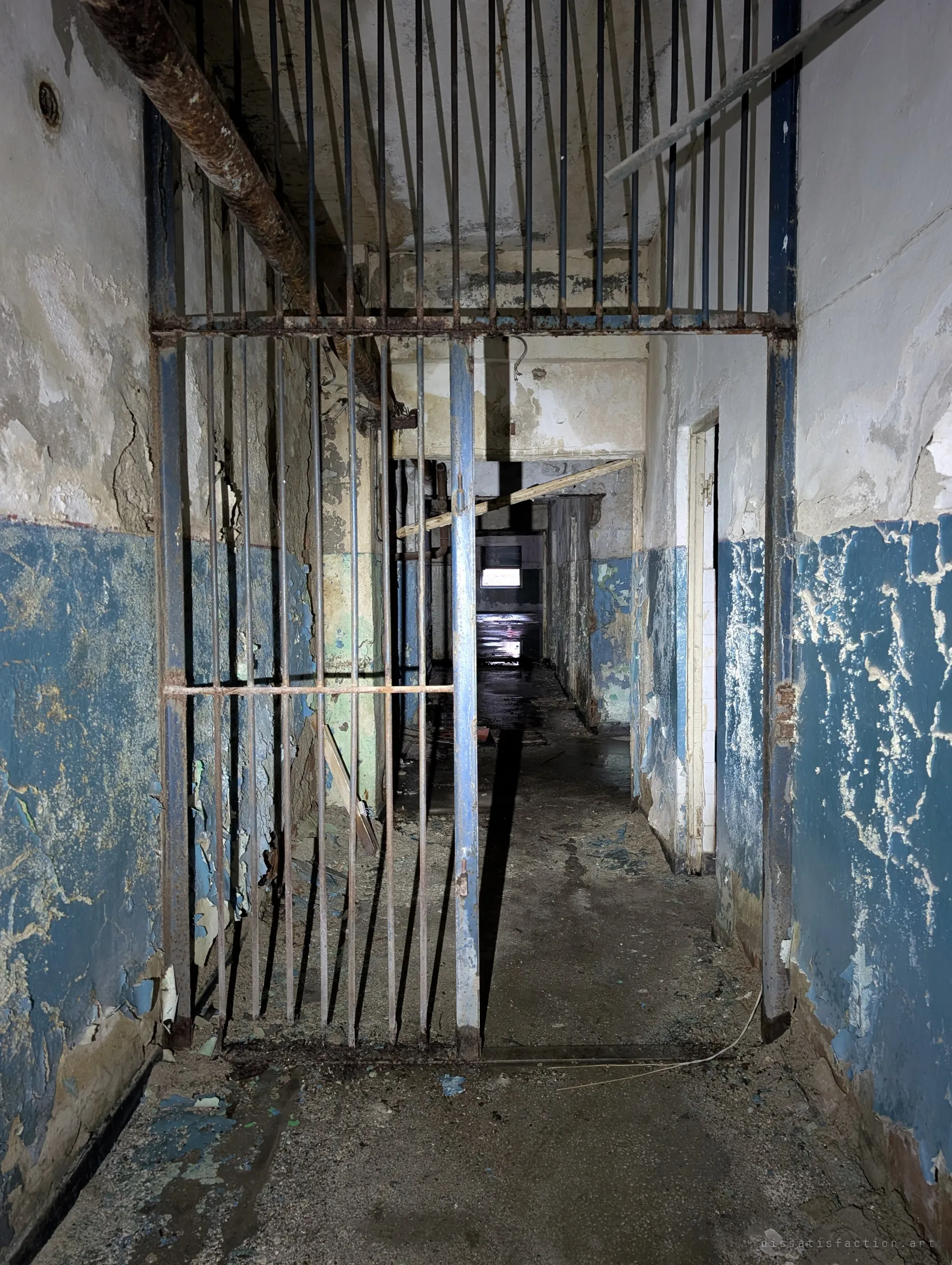 A barred steel gate stands open across a blue-painted corridor that runs deep into the building, wet floor glinting at the far end.