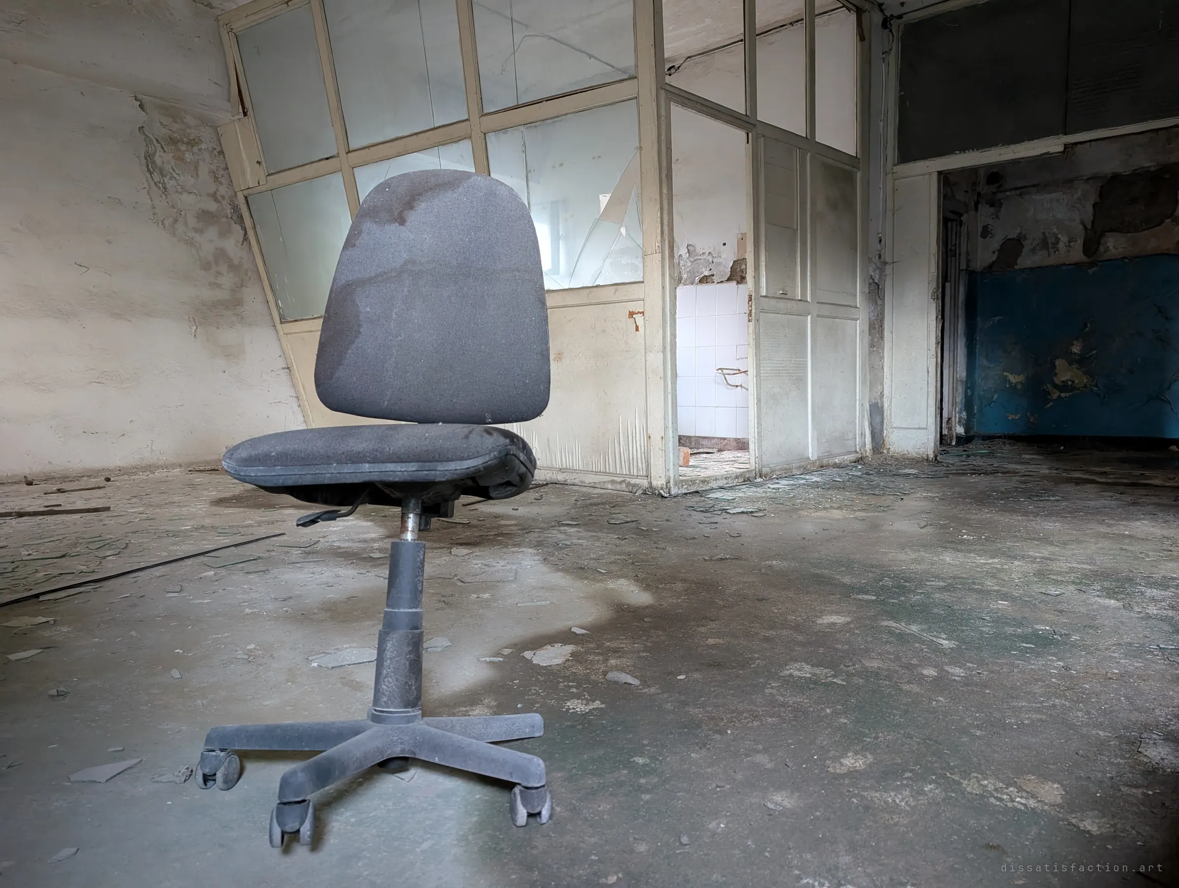 A lone office chair sits in an empty room in front of a glazed partition with shattered panes, blue paint flaking in the next room.