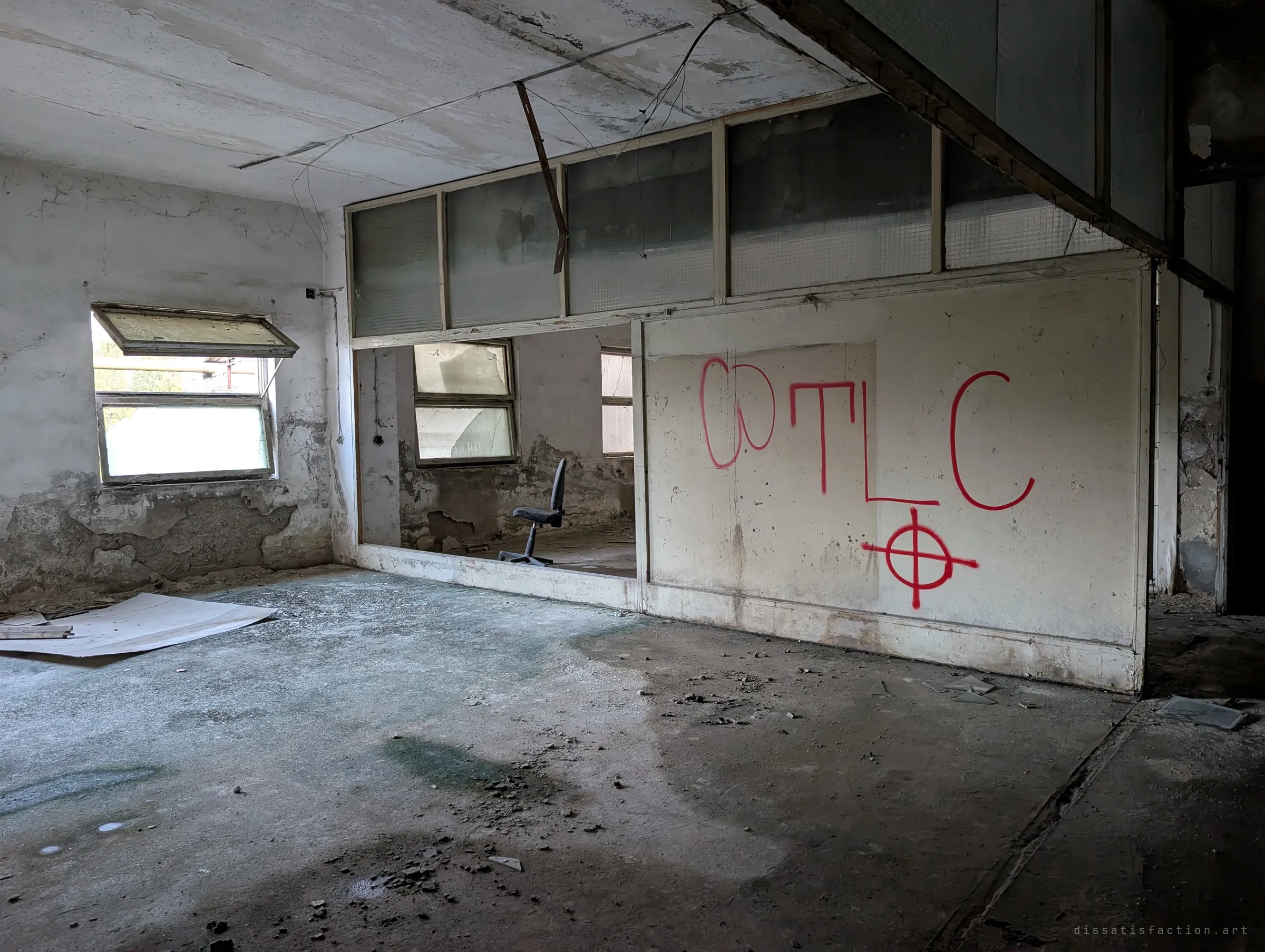 An abandoned open-plan office with a glazed partition and a single chair, red spray-painted letters and a crosshair on the wall.