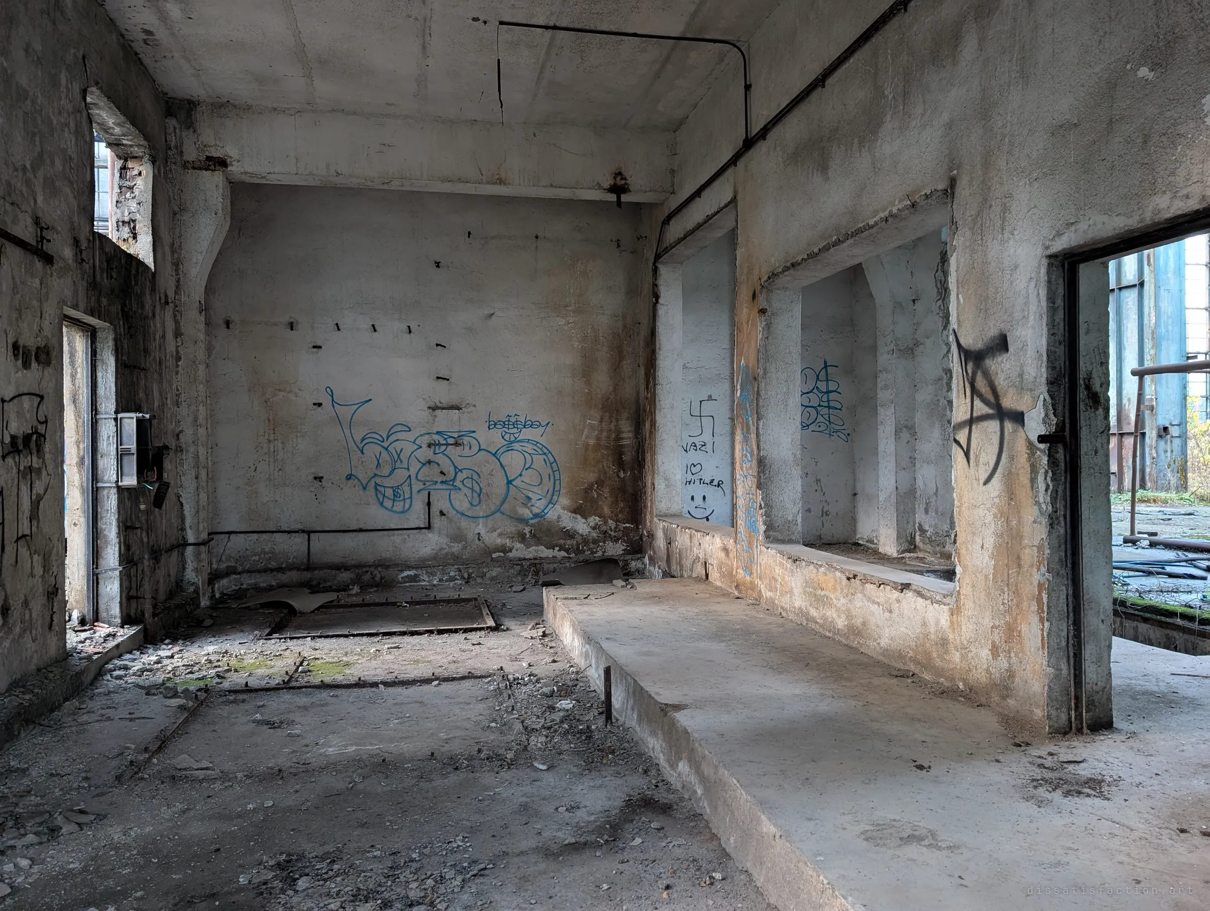 A raw concrete room with a raised plinth and floor frames where machinery once stood, walls covered in graffiti; a doorway opens onto the plant yard.