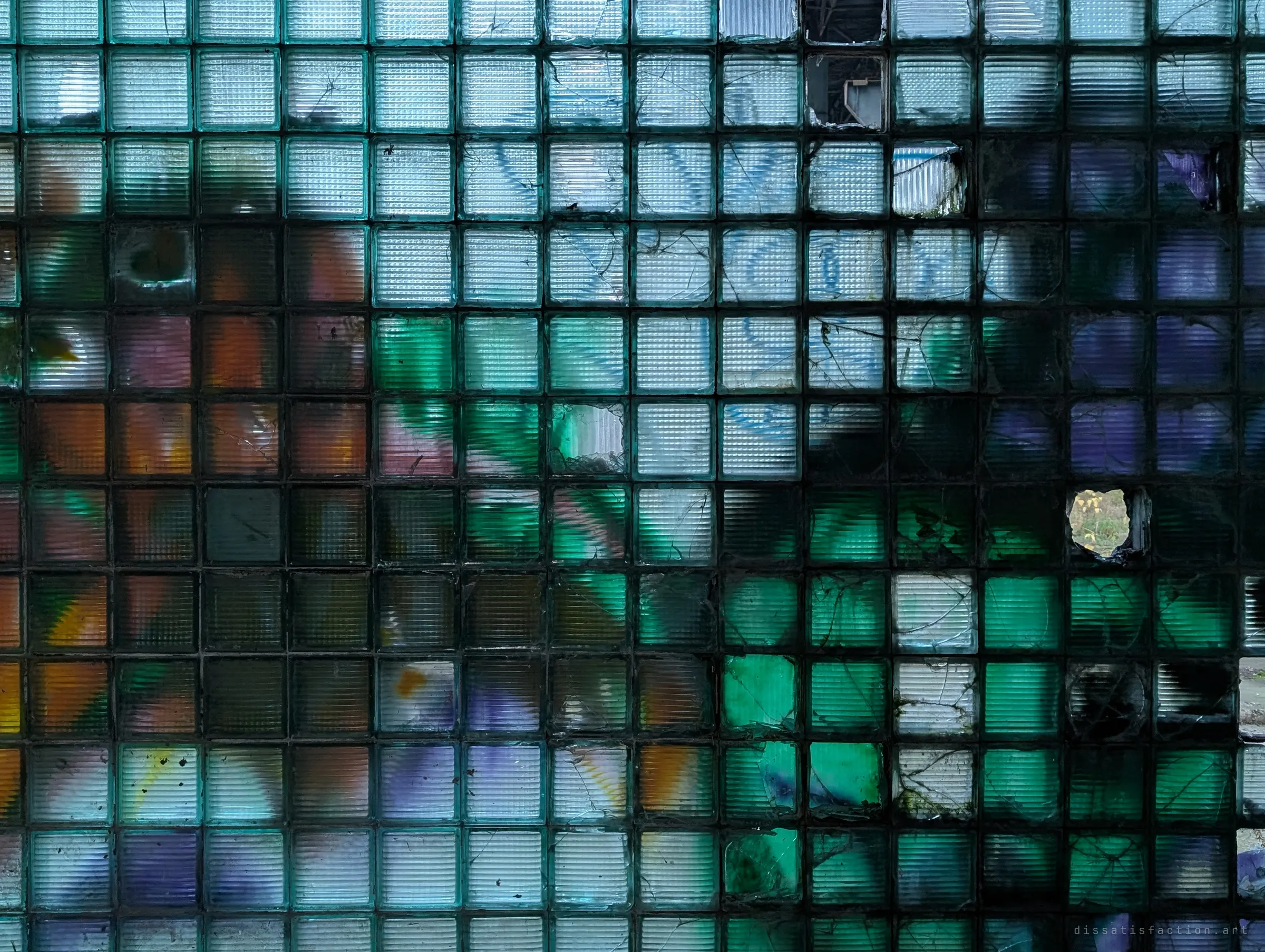 A wall of glass blocks glowing green, orange and violet from graffiti sprayed on the other side, a few blocks punched through.
