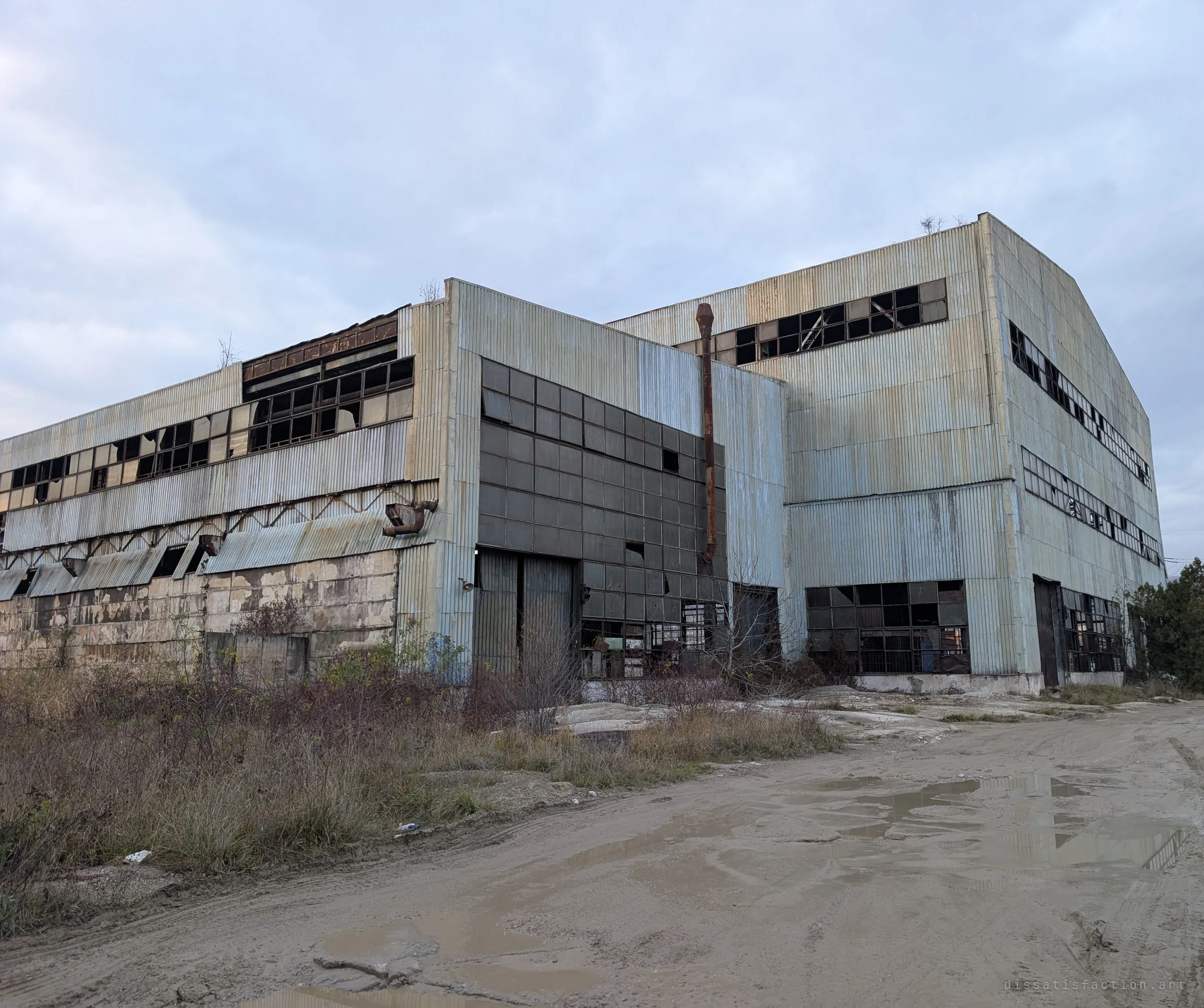 A long corrugated-steel factory hall with rows of smashed windows and a rusted flue pipe stands over a muddy, puddled yard under a grey sky. It shows the sheer scale of the abandoned works from outside.