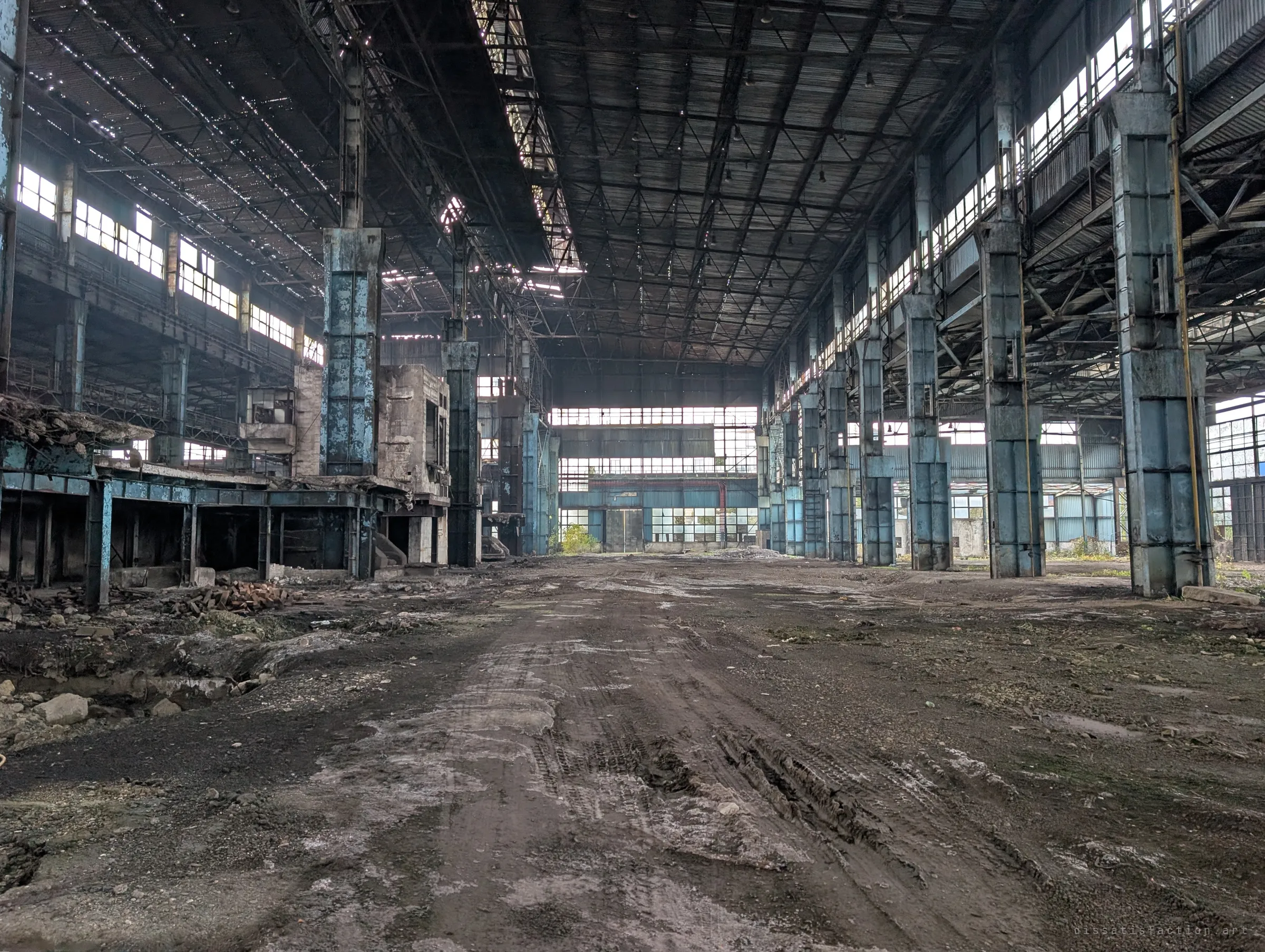 A vast, empty production hall stretches into the distance between rows of peeling blue steel columns, its roof pierced with pinholes of daylight. Tyre tracks in the dirt floor mark where machinery was hauled out.