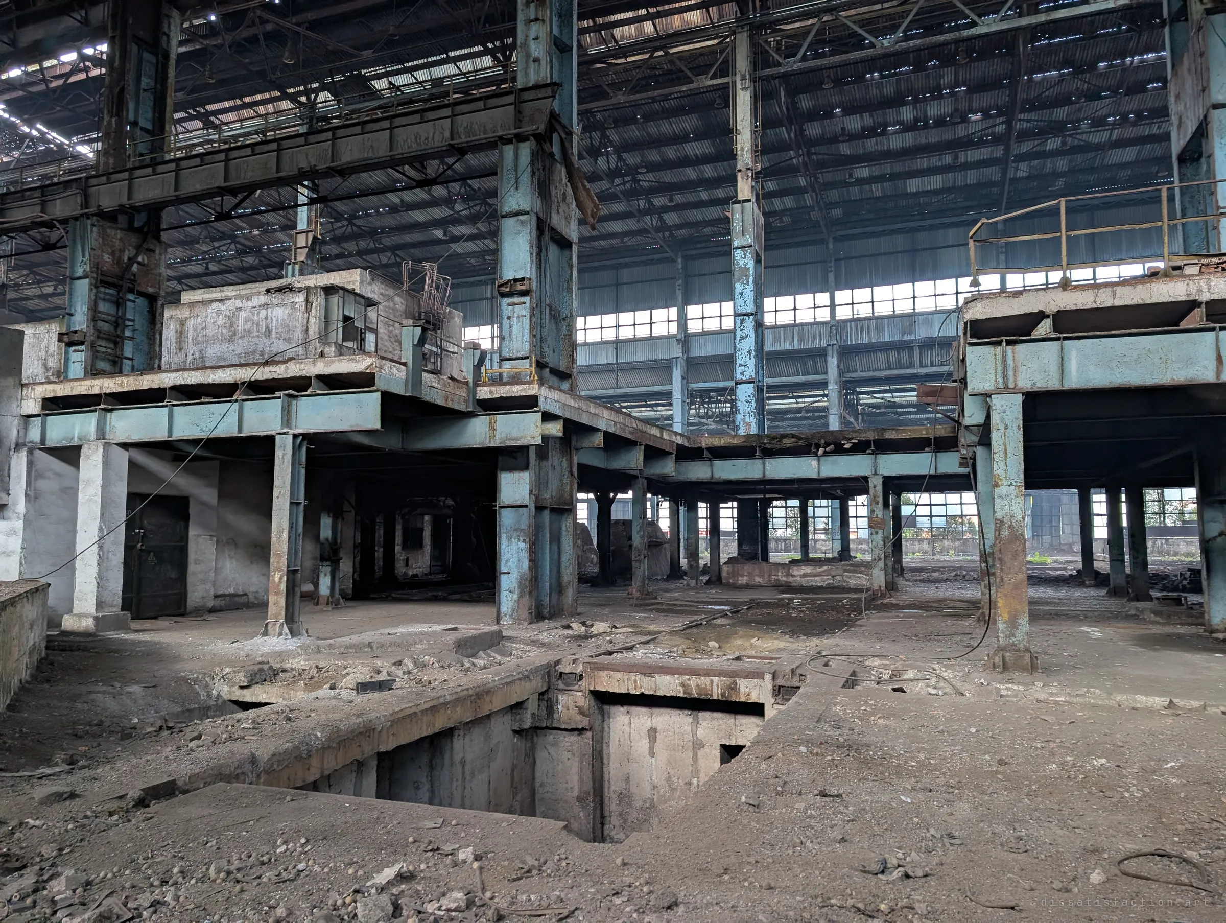 Blue steel platforms and walkways hang over an open concrete pit in the floor of a huge hall, with light slanting in through a doorway on the left. The foundations of long-removed heavy machines are laid bare.