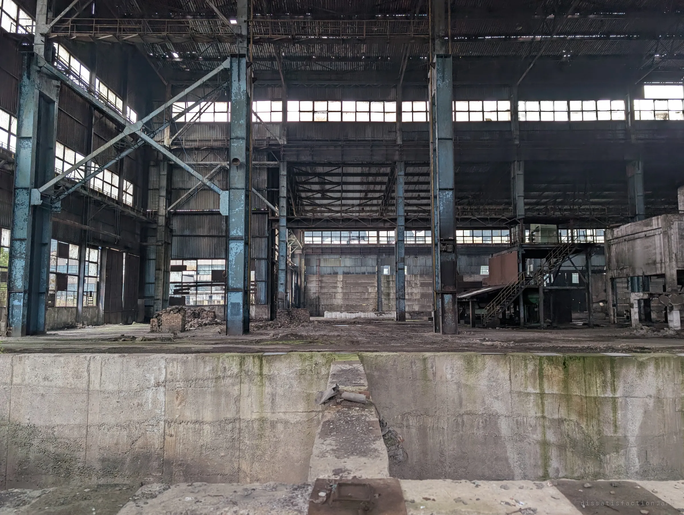 Seen across the edge of a deep concrete pit, the hall opens up into tall blue columns, cross-bracing and a long band of high windows. A staircase climbs to a small steel structure on the right.