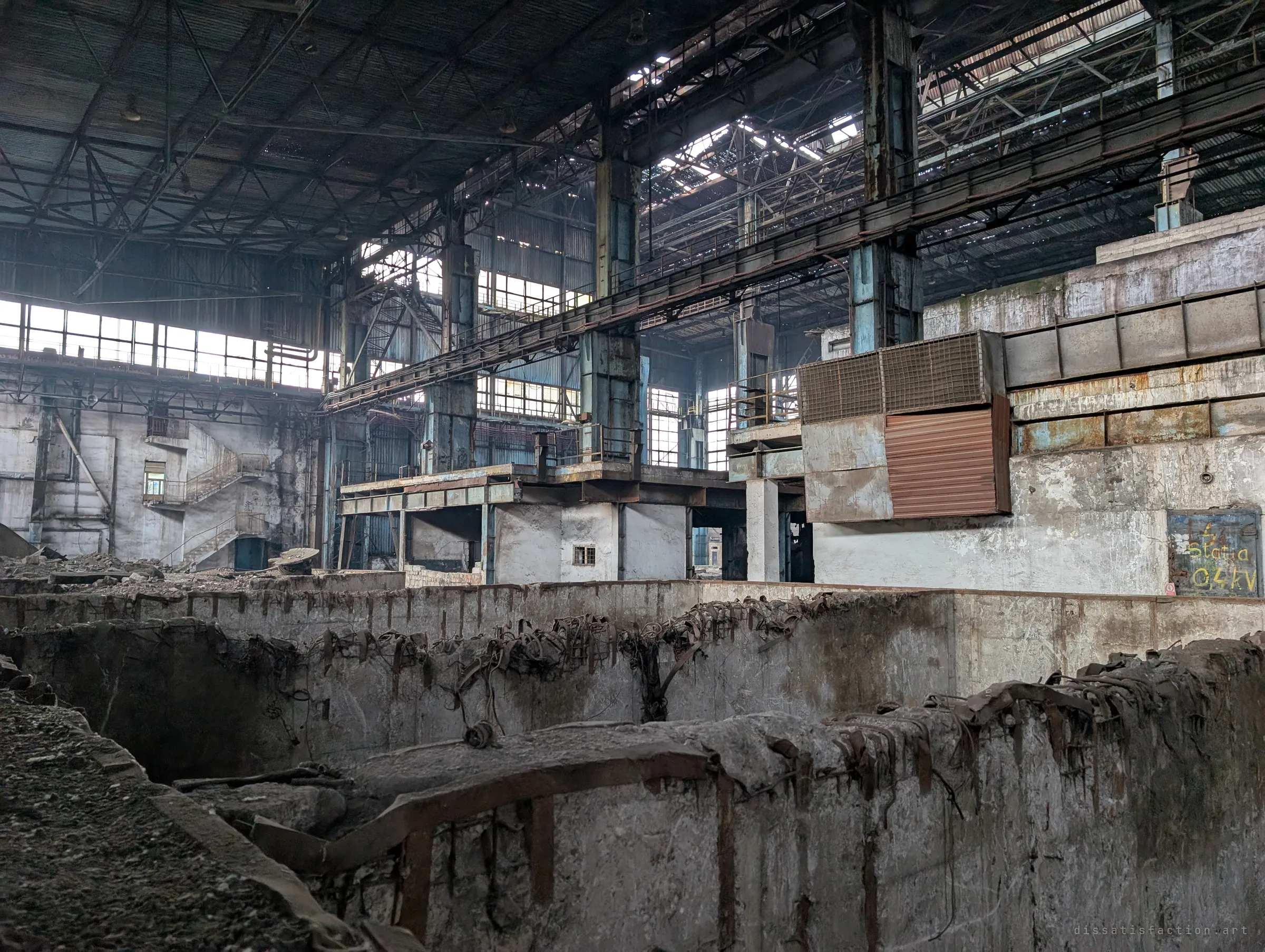 Twisted steel strapping curls over the rim of a ruined concrete pit, while behind it platforms, a walkway and a hand-painted electrical substation door fill the hall. Industrial layers stacked on top of each other.