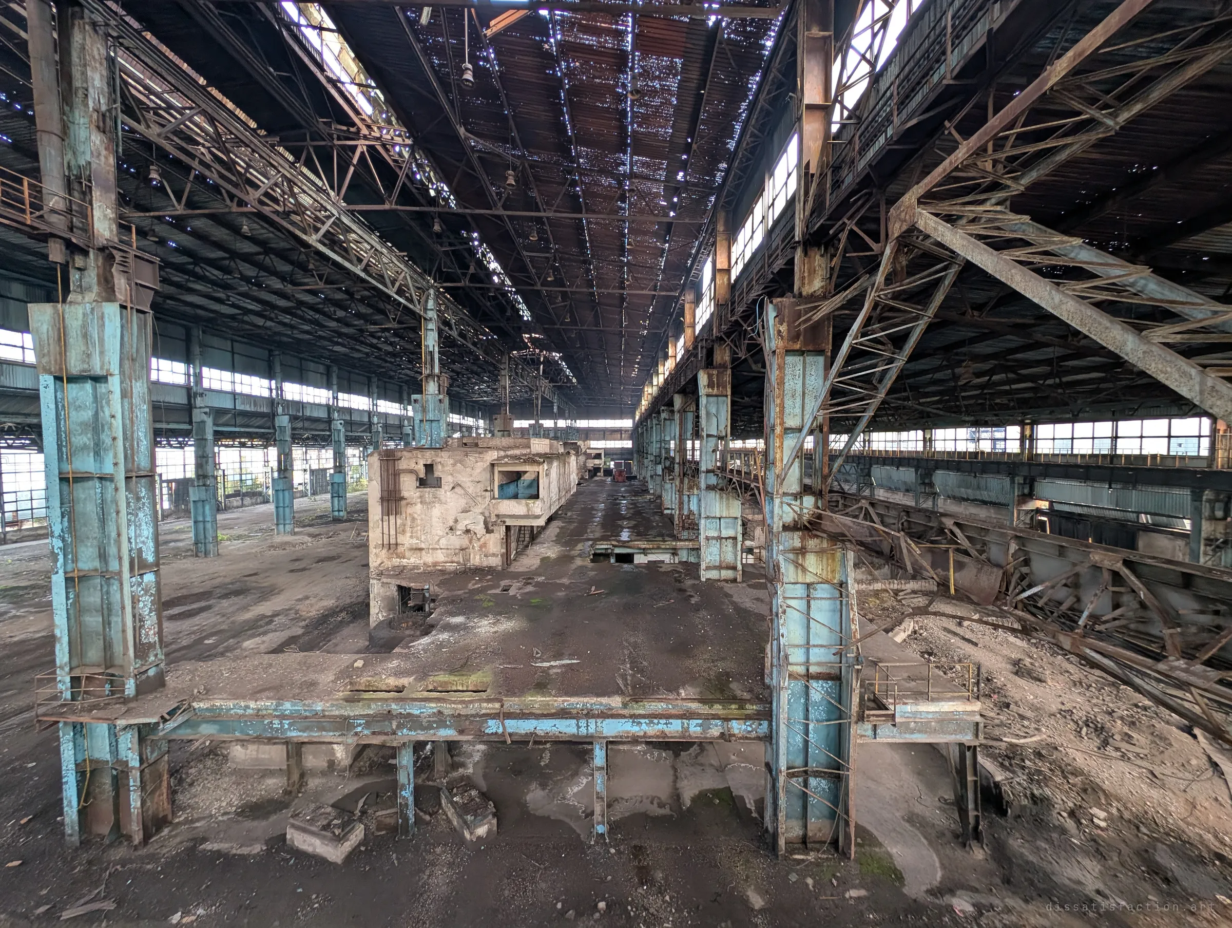From a raised vantage point, a long bay of the hall recedes between rust-streaked blue columns and a skeletal roof full of holes. A concrete control cabin sits in the middle of the emptied floor.