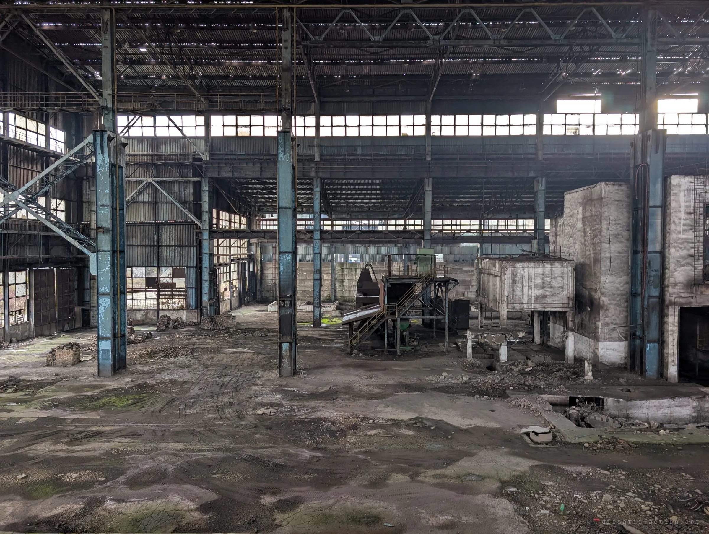 An elevated view over a wide hall floor scattered with rubble and brick piles, with a small steel staircase and platform standing alone among blue columns. Daylight pours through the continuous window band above.