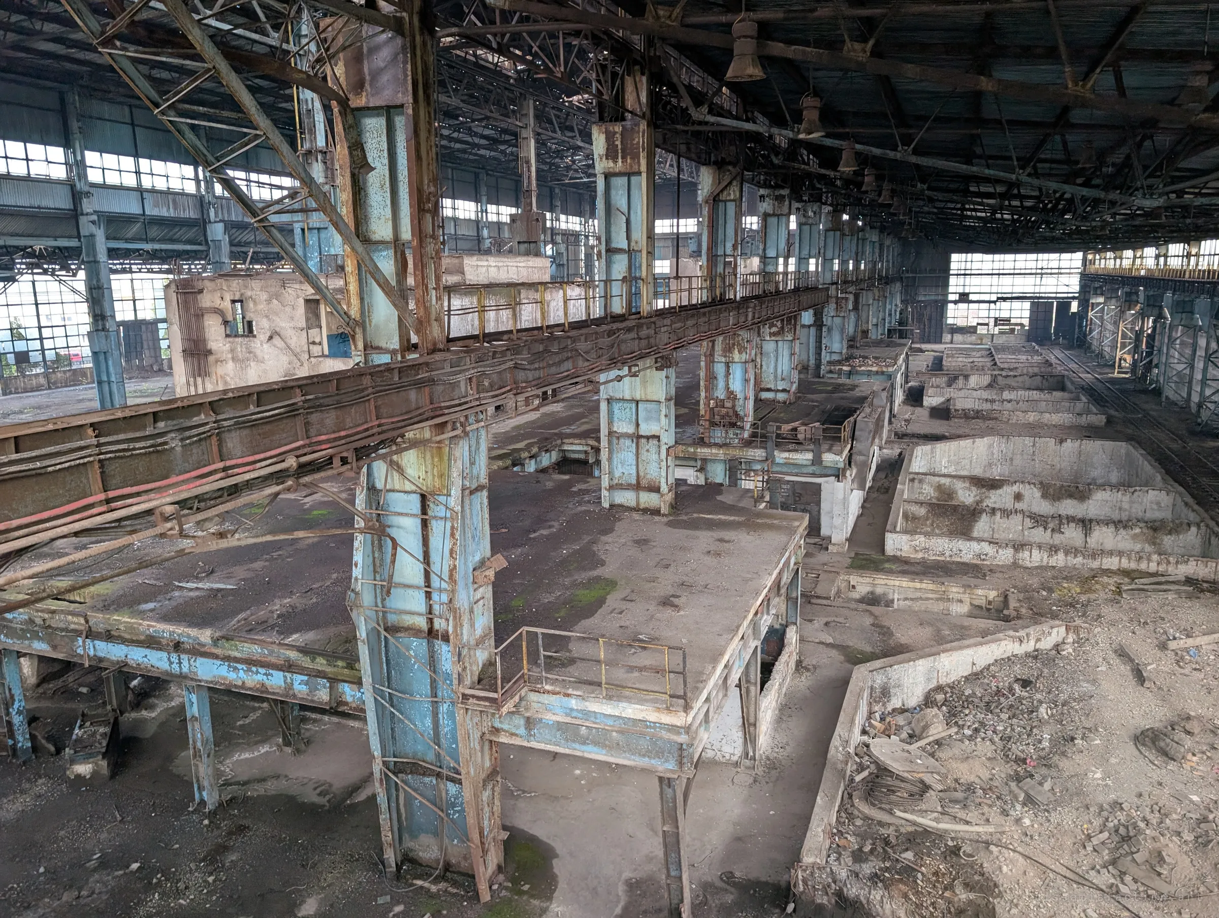 Looking down from a crane-rail level over cable trays, rusted blue platforms and a row of empty concrete pits running the length of the hall. The layout reveals how heavy work once moved through the building.