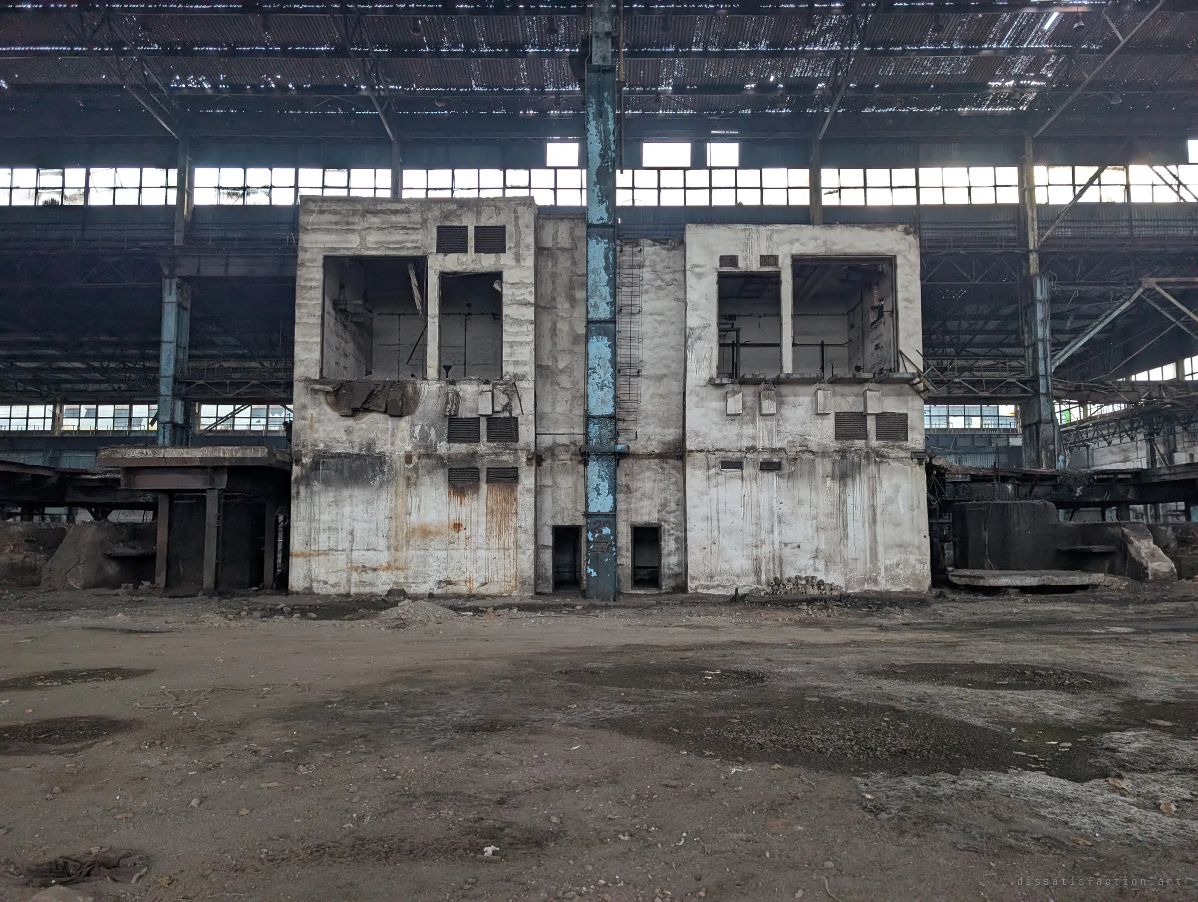 A two-storey concrete control building, its windows gaping and walls stained with rust, stands alone on the dirt floor of the hall beneath a perforated roof. A lone blue column rises straight through its middle.
