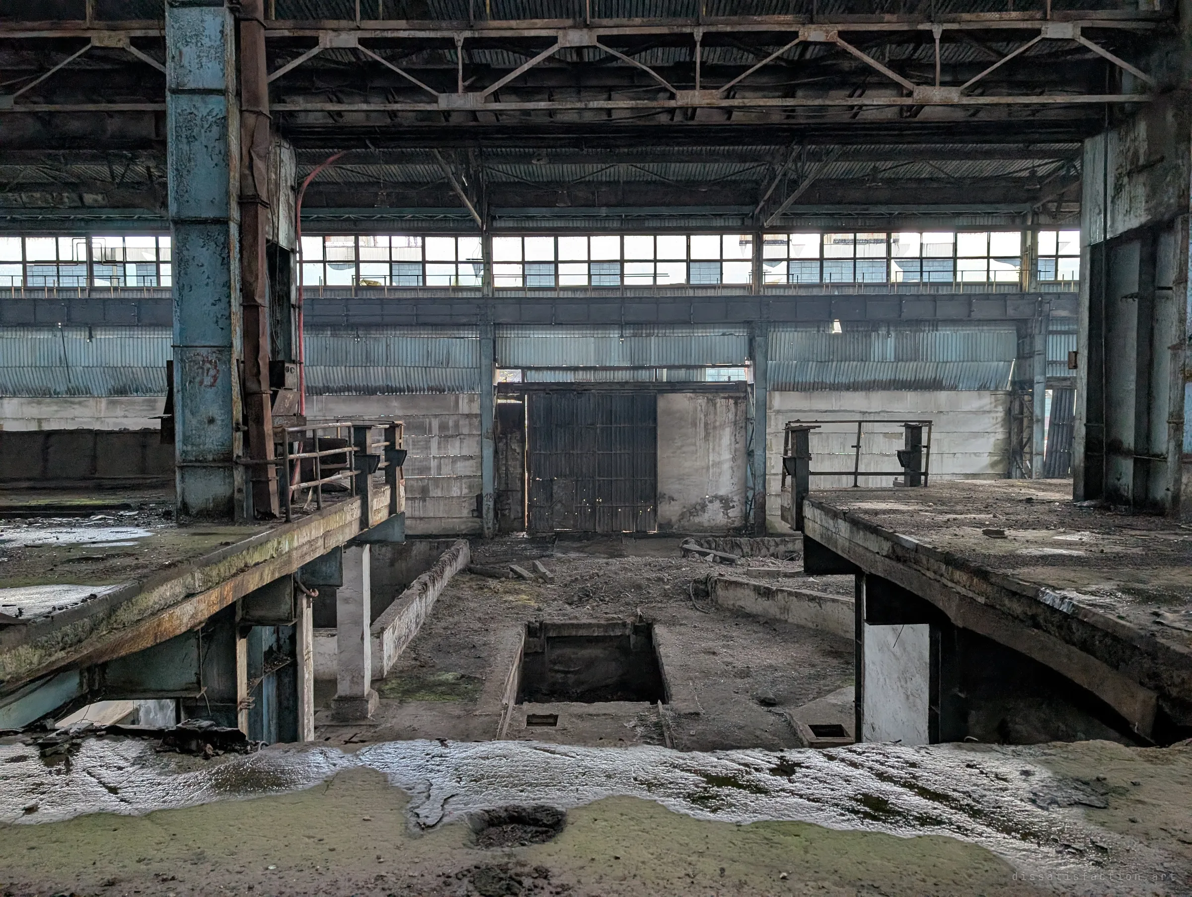 Between two broken concrete decks, a trench and a small pit lead the eye to a tall iron gate in the far wall of the hall. Wet, mossy concrete in the foreground glistens in the soft light.