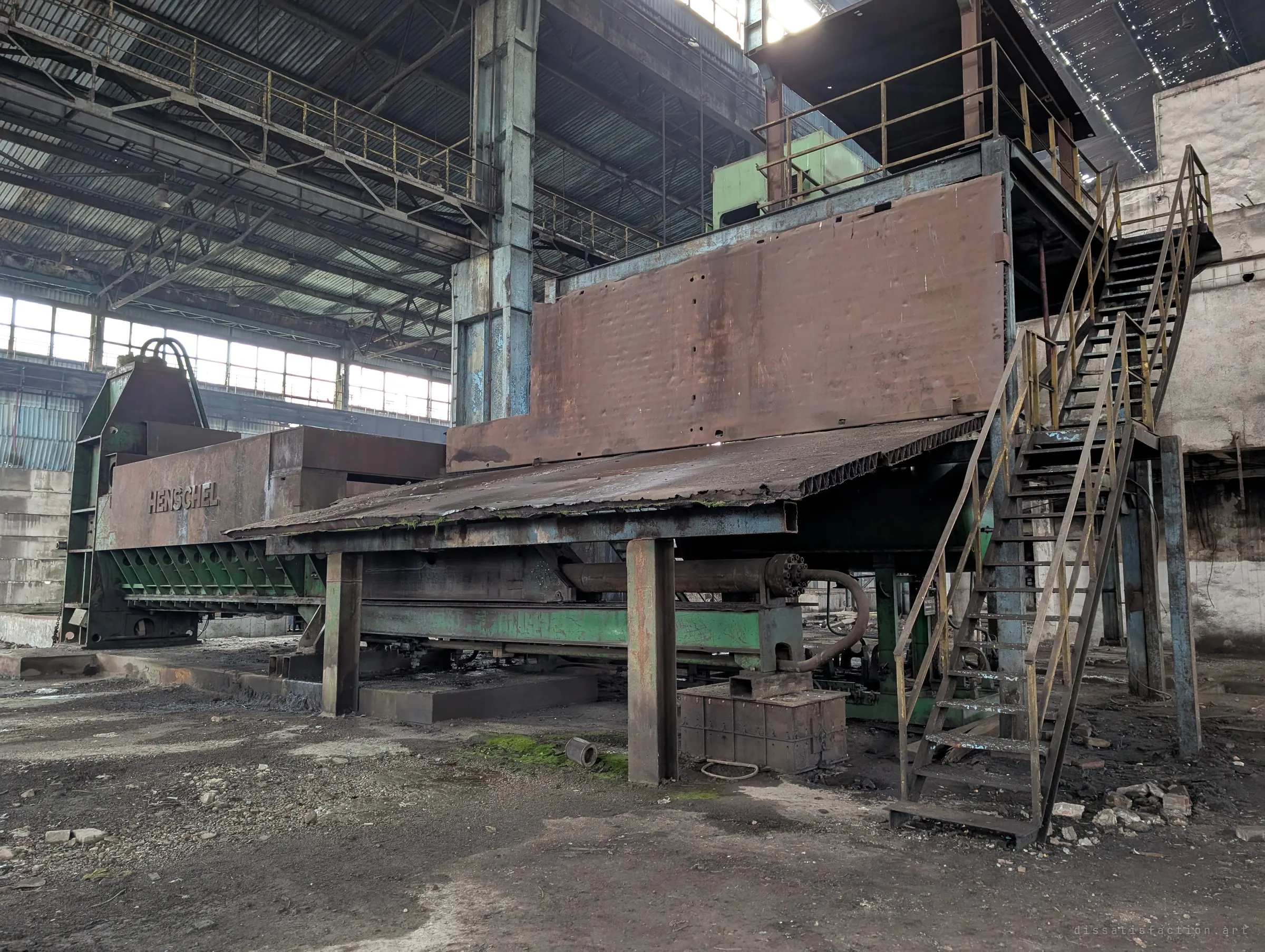 A huge rusted press with a green frame and steel stairs to its upper deck stands in a hangar-sized hall under a broken roof. Its scale shows the heavy machinery this plant was built around.