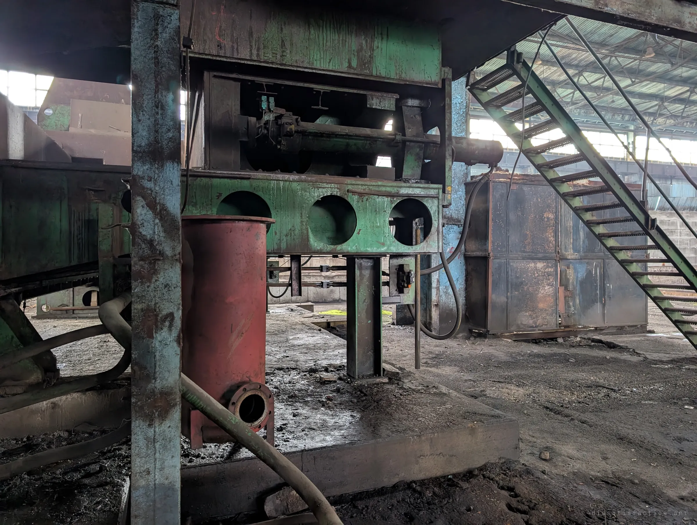 Underneath a green press frame with round cut-outs, a red cylinder and loose hydraulic hoses sit on an oil-stained plinth. A green stair climbs away to the right into the bright hall.