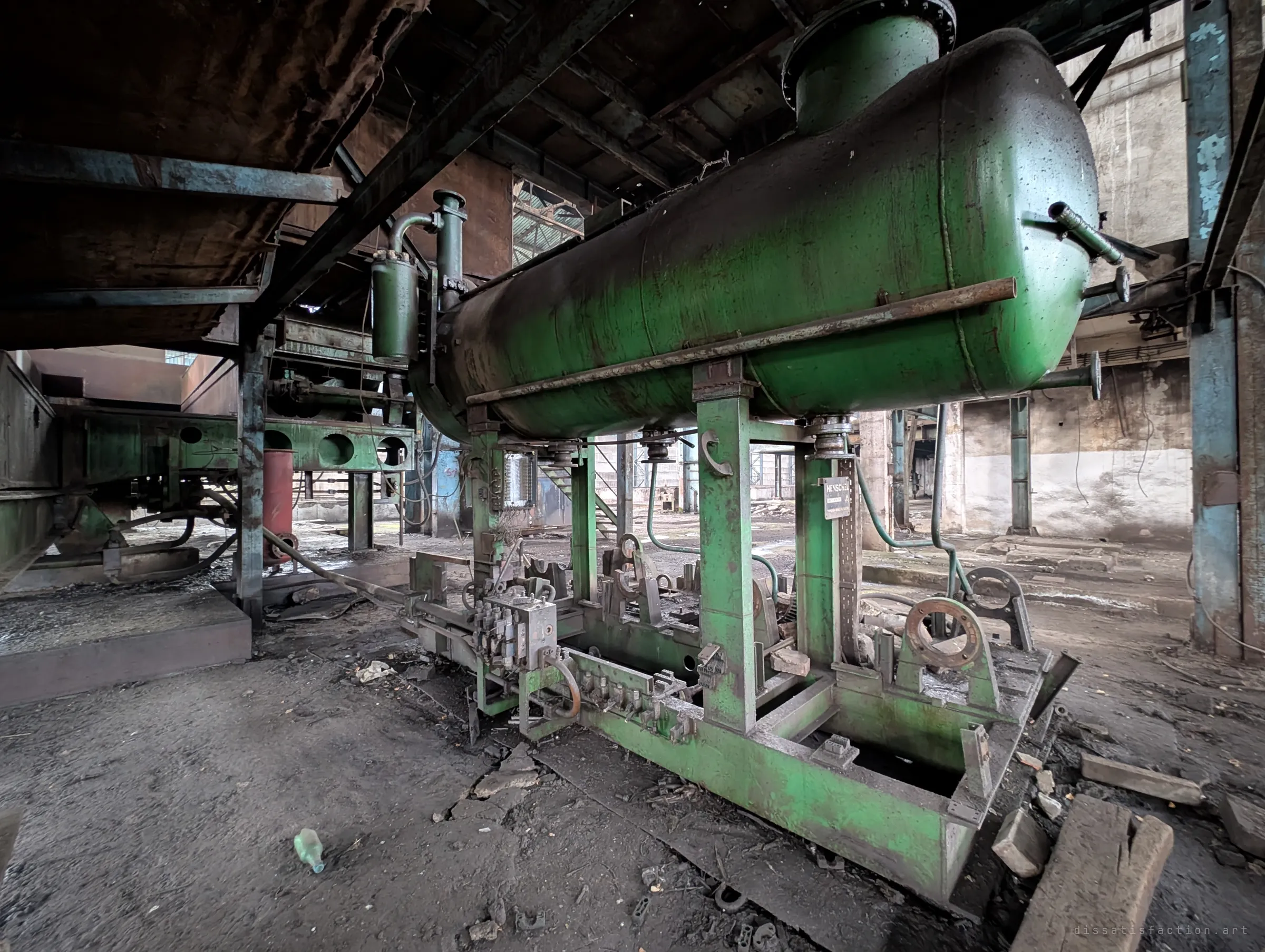 A wide view of a green pressure tank mounted on a frame of valves and pipe clamps beneath the press gantry. Rubble and broken bottles litter the floor around it.