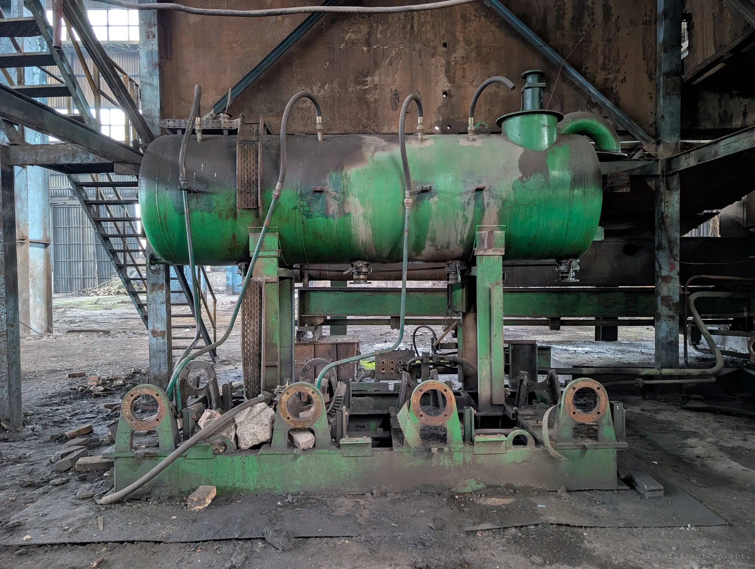 The green tank seen straight on, with hoses looping into its top and rusted round mounts along the base. It sits against the rusted wall of the press, stairs rising on the left.