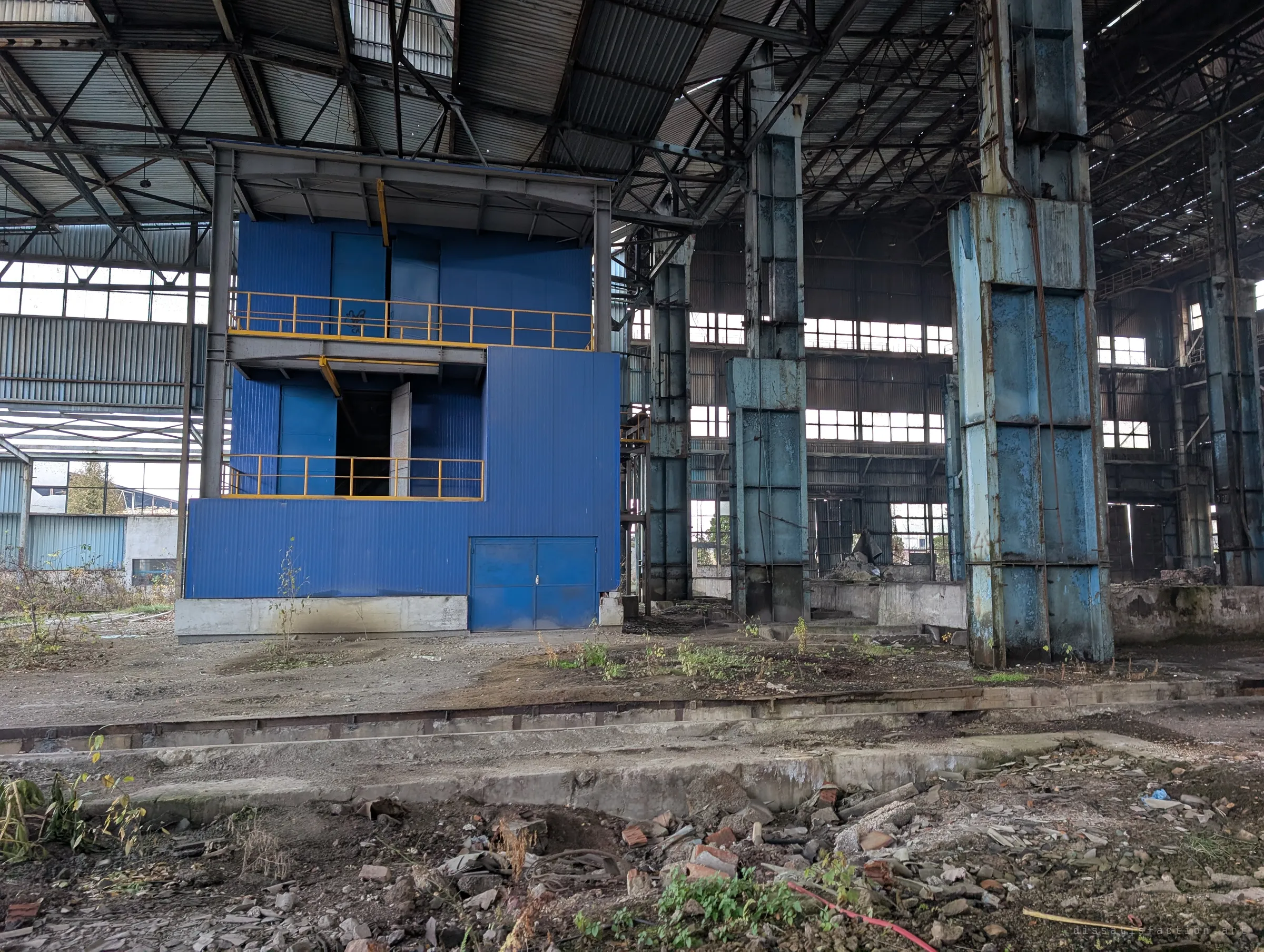 A two-storey blue control box with yellow railings stands in the hall beside massive pale-blue crane columns. Rows of windows behind light the long empty building.