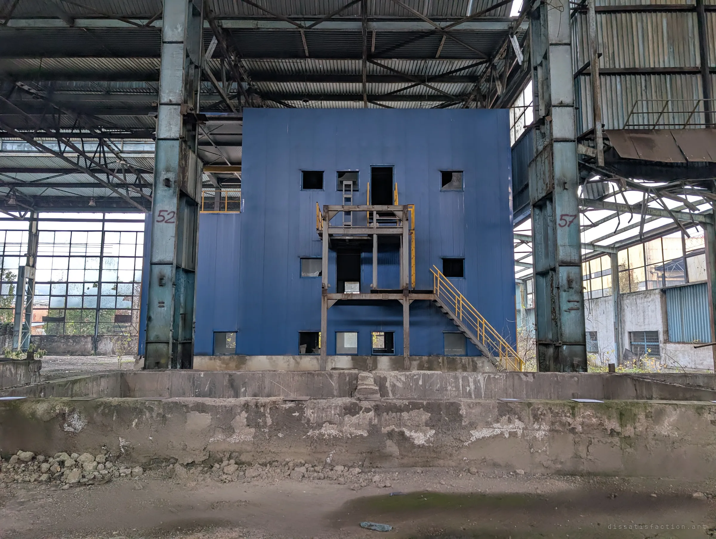 The blue block seen head-on across an empty concrete pit, framed by two numbered steel columns. Small square windows and a yellow stair give it the look of a face.