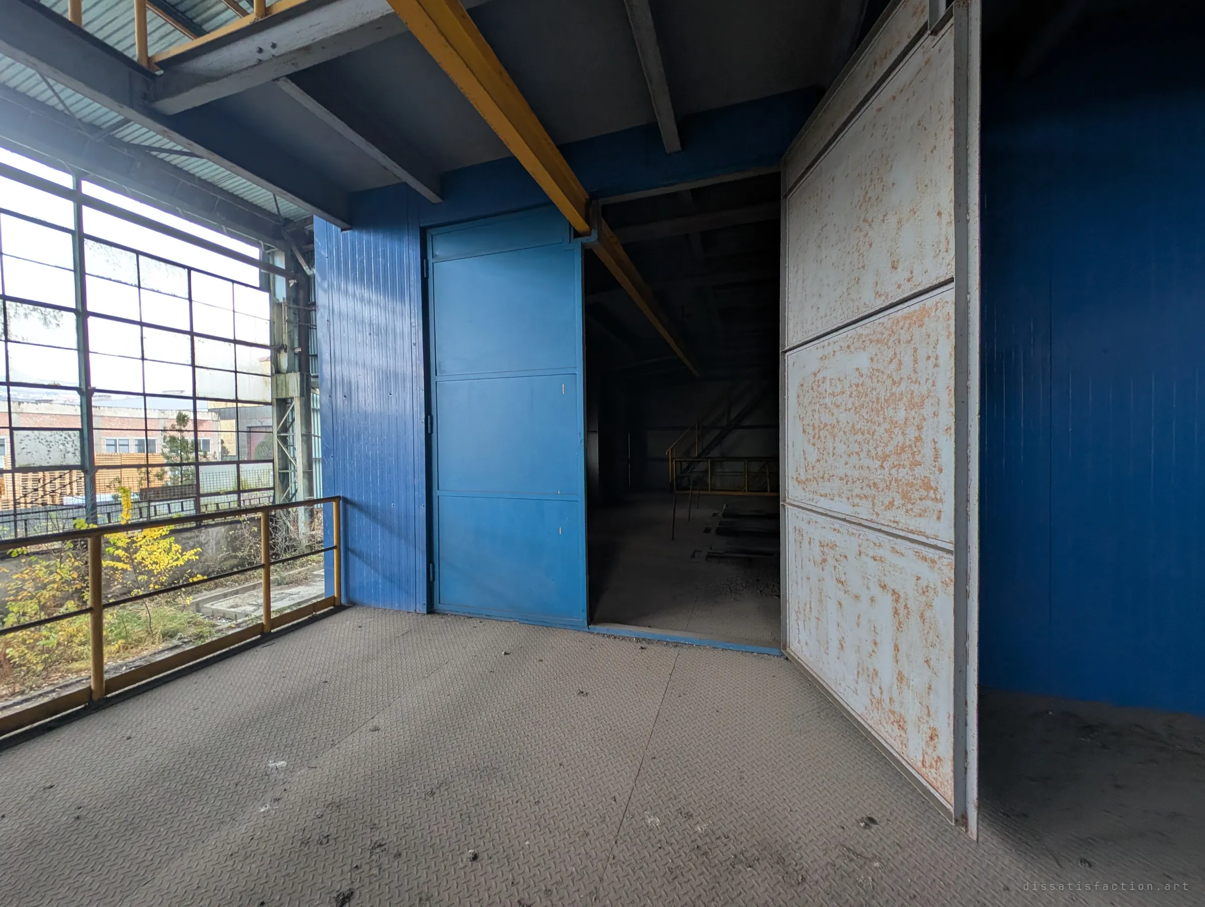 On the upper landing of the blue block, a pale door hangs open onto a dark room with a yellow crane rail overhead. A tall grid window lets in daylight from the yard.