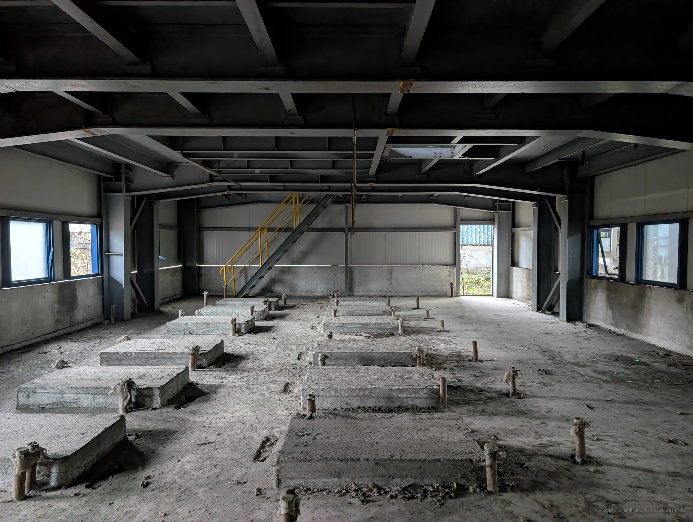 A dim room of bare concrete machine plinths with pipe stubs sticking up in rows, a yellow stair at the far end and a door open to the outside. Whatever stood here has been removed.