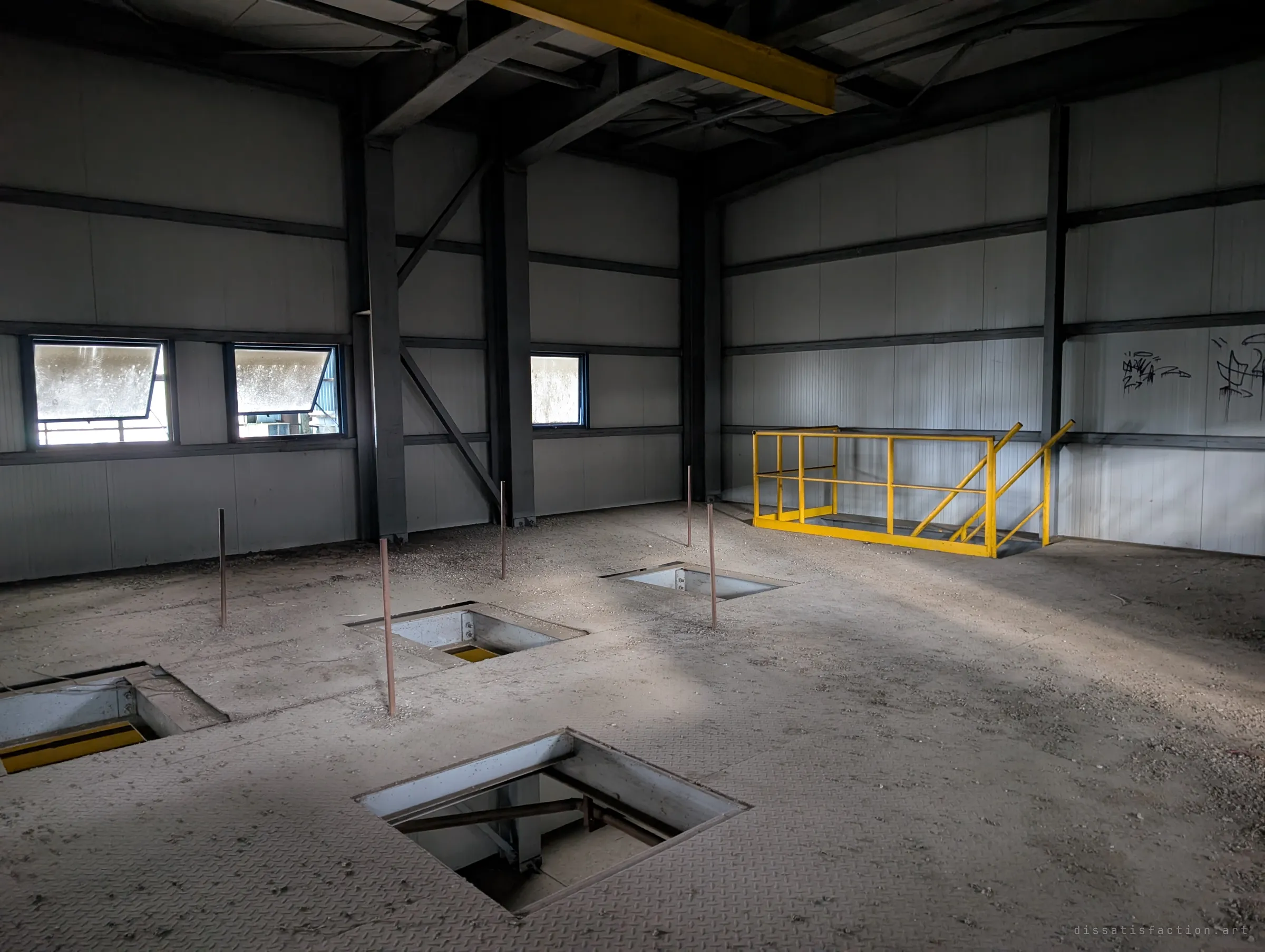 An upper floor with square hatches cut into the checker-plate floor, thin rods standing up and a yellow railing by the far wall. Light from small windows falls across the dust.