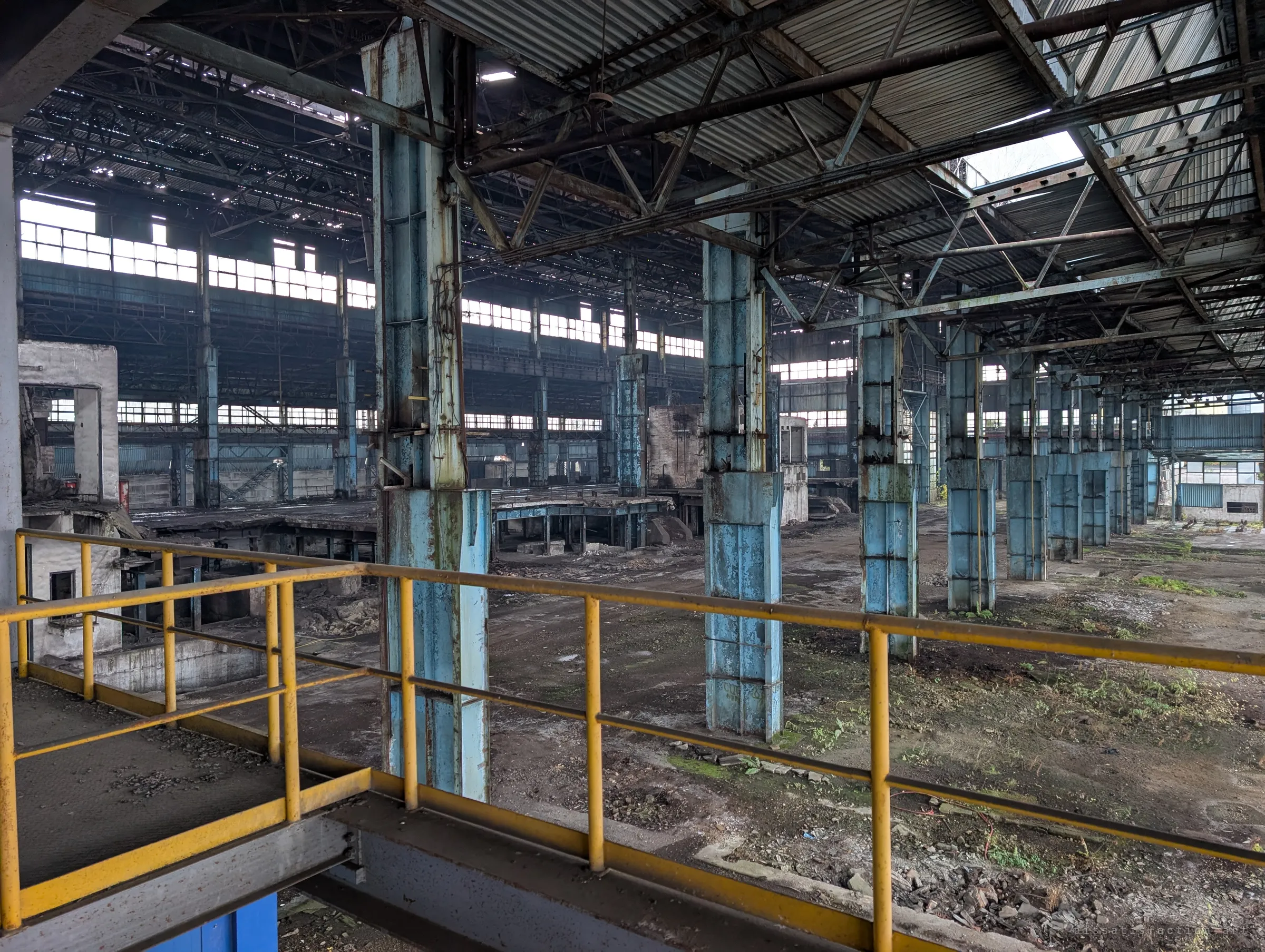 From a yellow-railed platform, rows of flaking pale-blue columns run off into the vast hall, its floor gone to weeds and rubble. Broken windows along the far walls show how big the plant was.