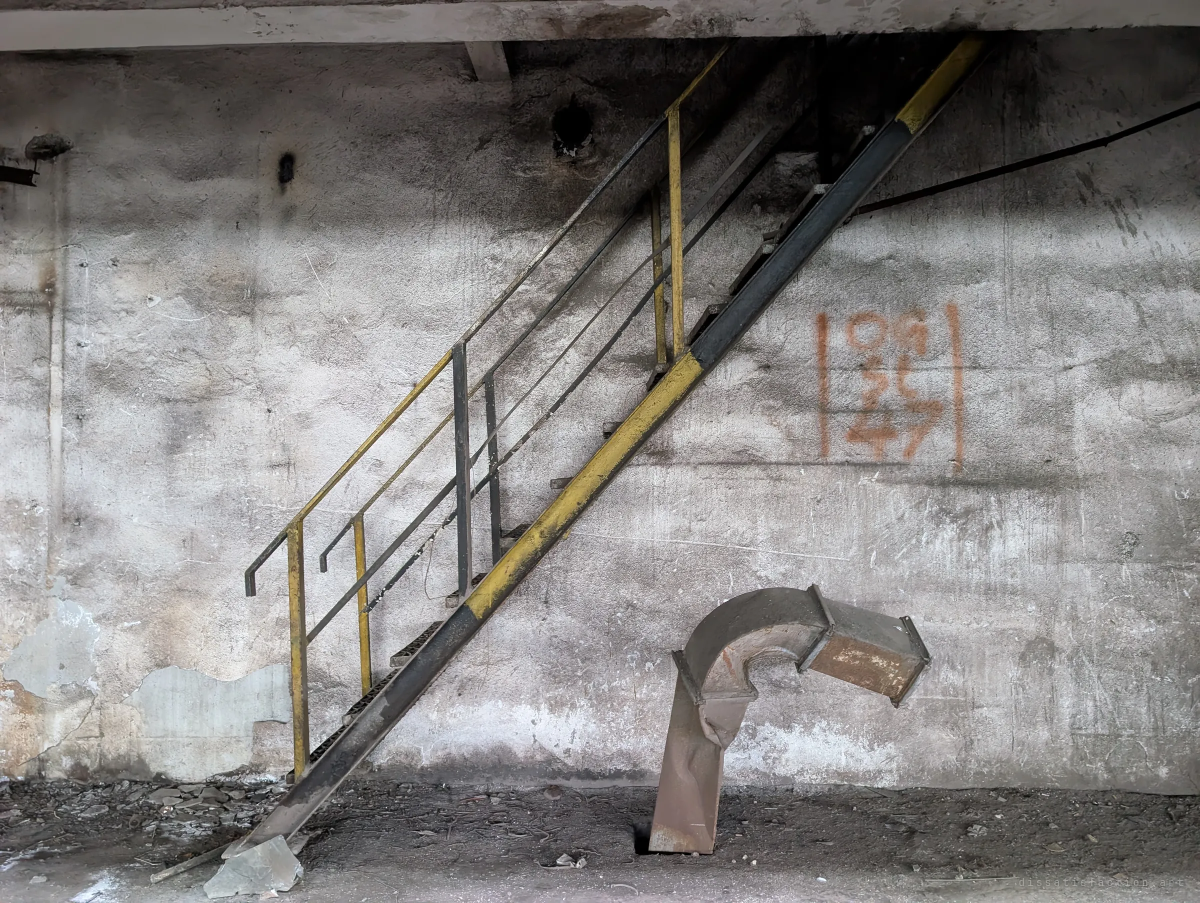 A yellow-and-black steel stair climbs a stained concrete wall beside a hand-painted column grid marker, with a bent duct stub leaning out of the floor.