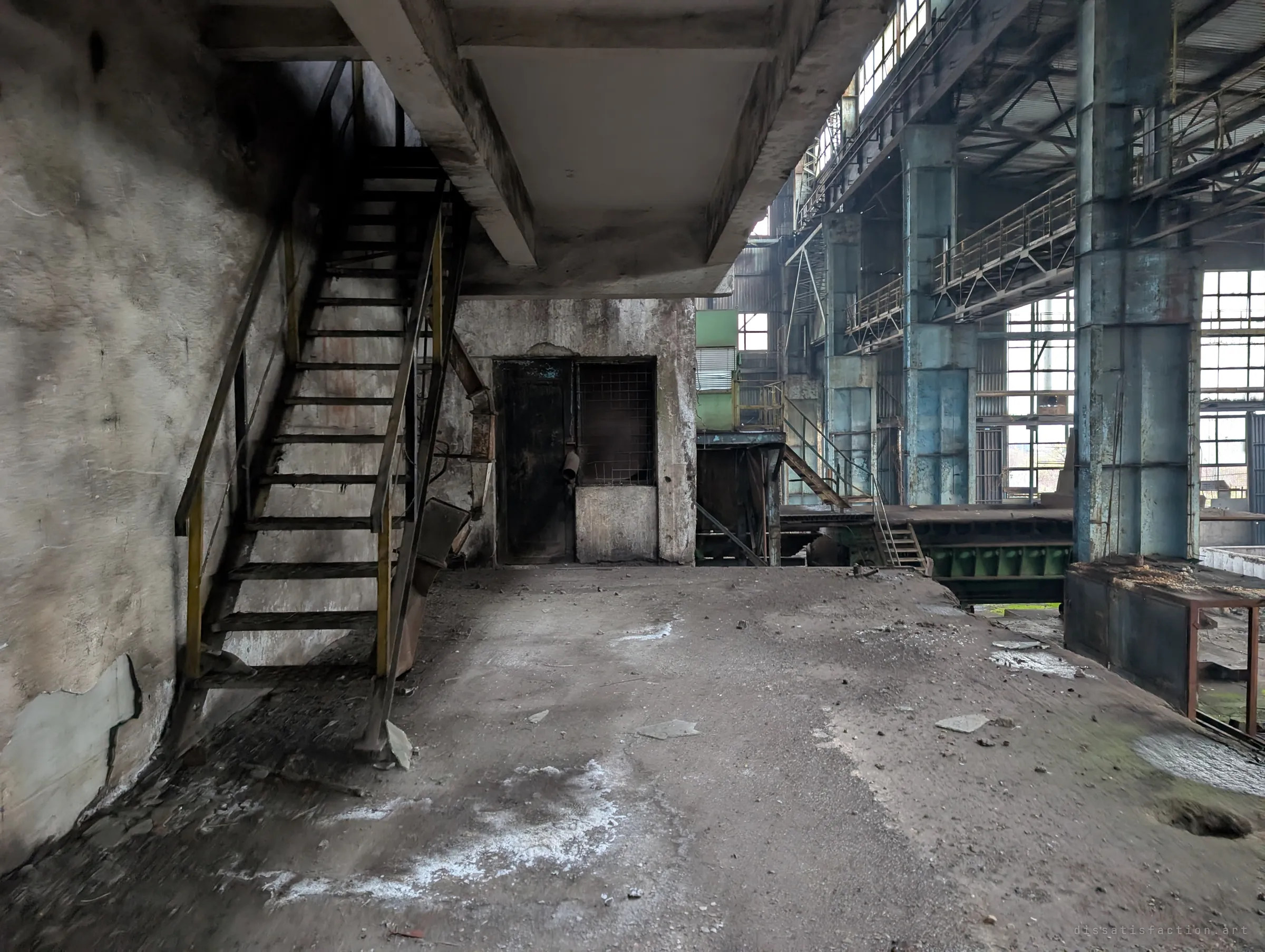Under a concrete landing, a steel stair and a sealed control booth open onto the main hall, where blue-painted columns and catwalks rise toward tall grid windows.