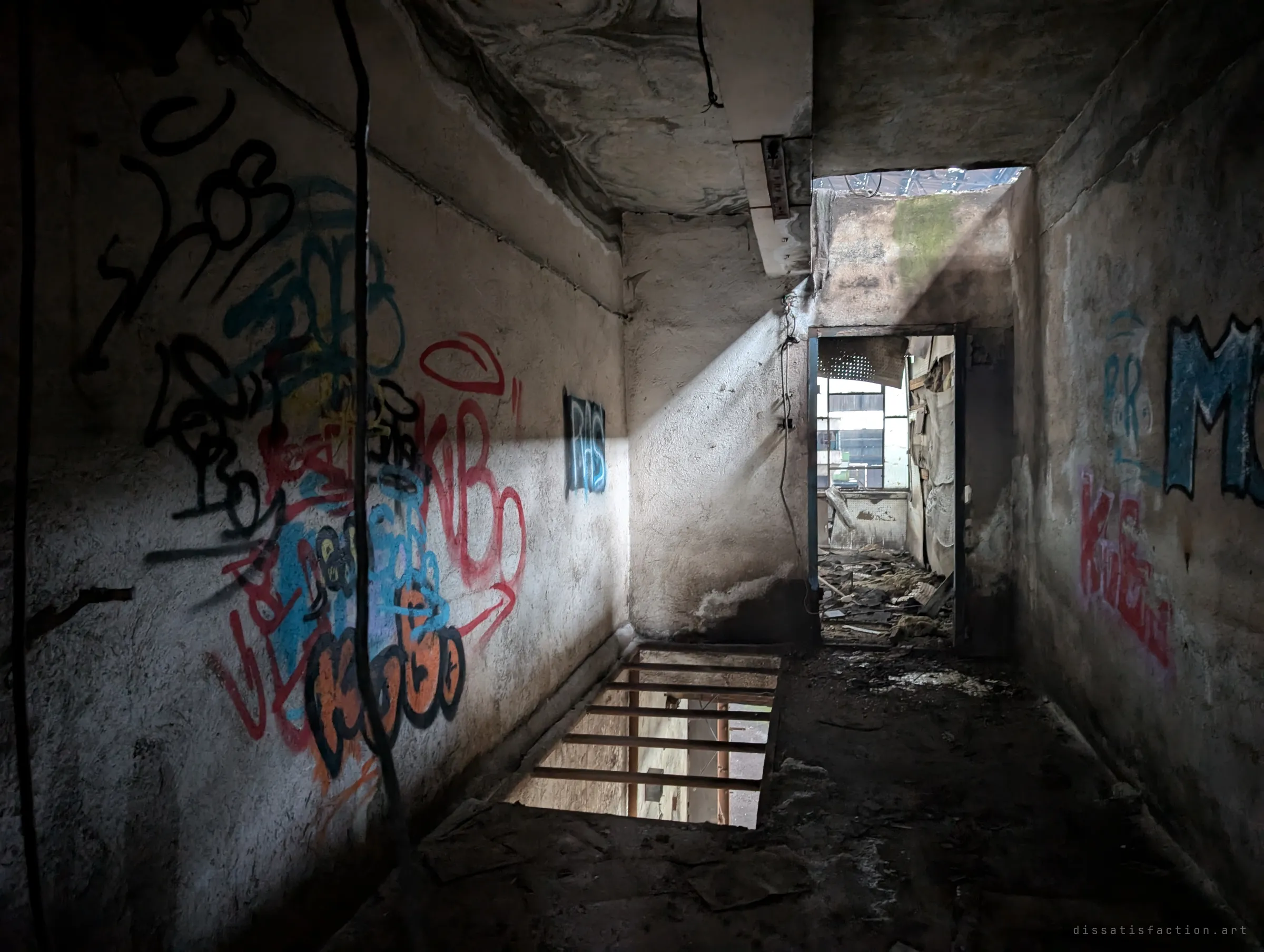 A graffiti-covered service corridor lit by a slanting beam of daylight, with an open floor grate and a doorway into a wrecked room beyond.