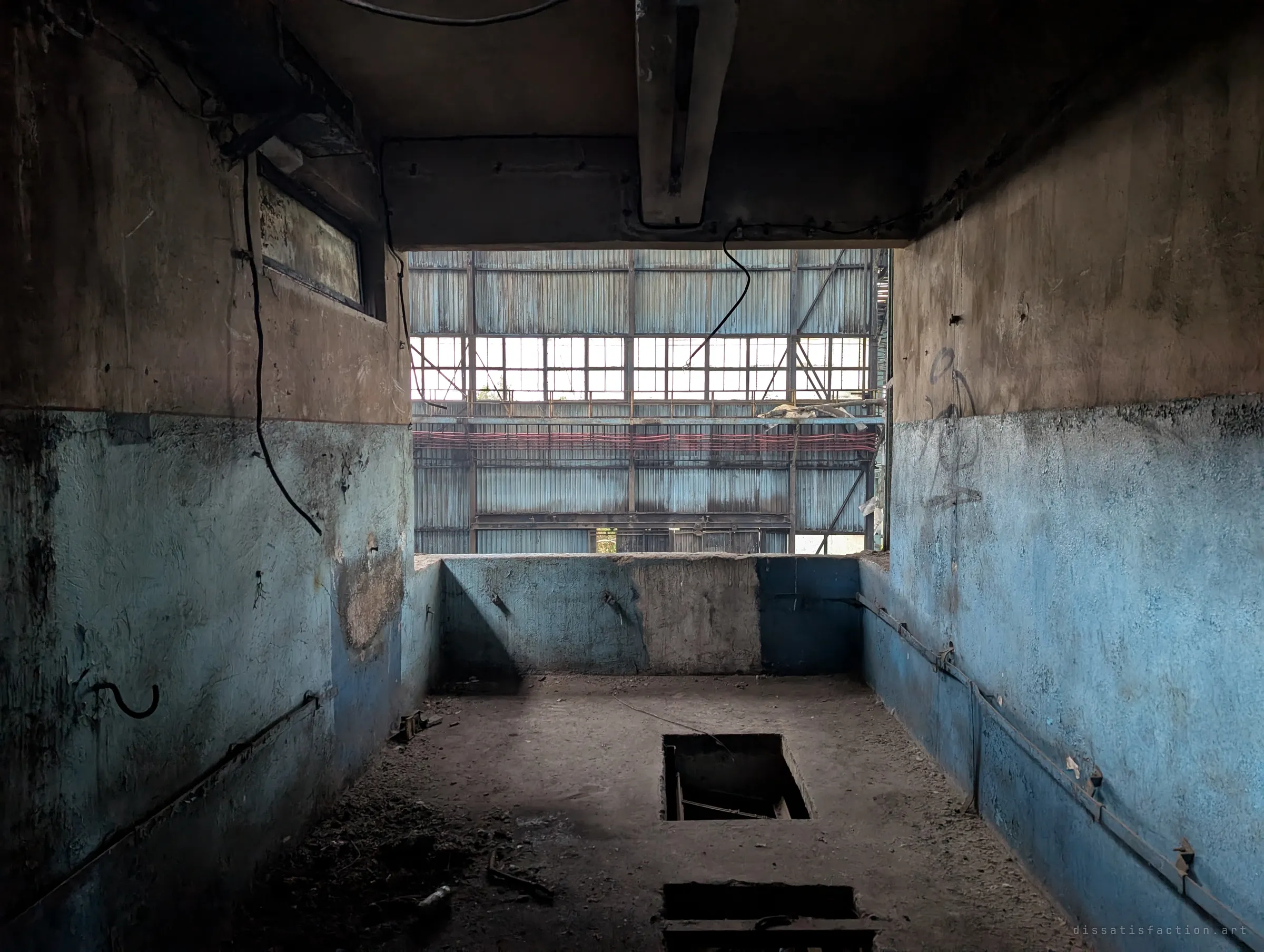 A two-tone blue and brown concrete room with open floor hatches looks out through a glassless opening onto the corrugated wall and window bands of the hall.