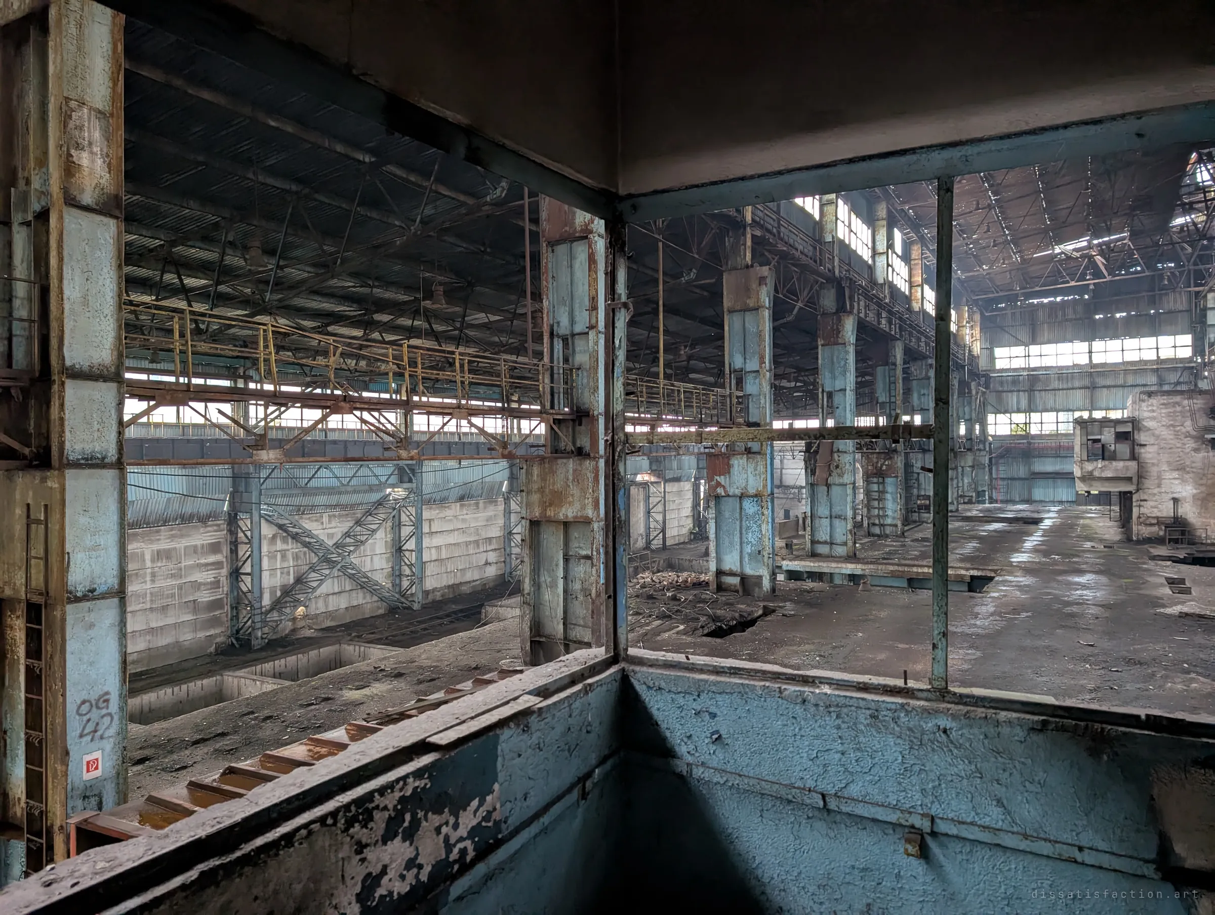 From a crane-operator style booth with empty window frames, the view sweeps across the vast hall: gantry walkways, rusting blue columns and a stripped concrete floor.