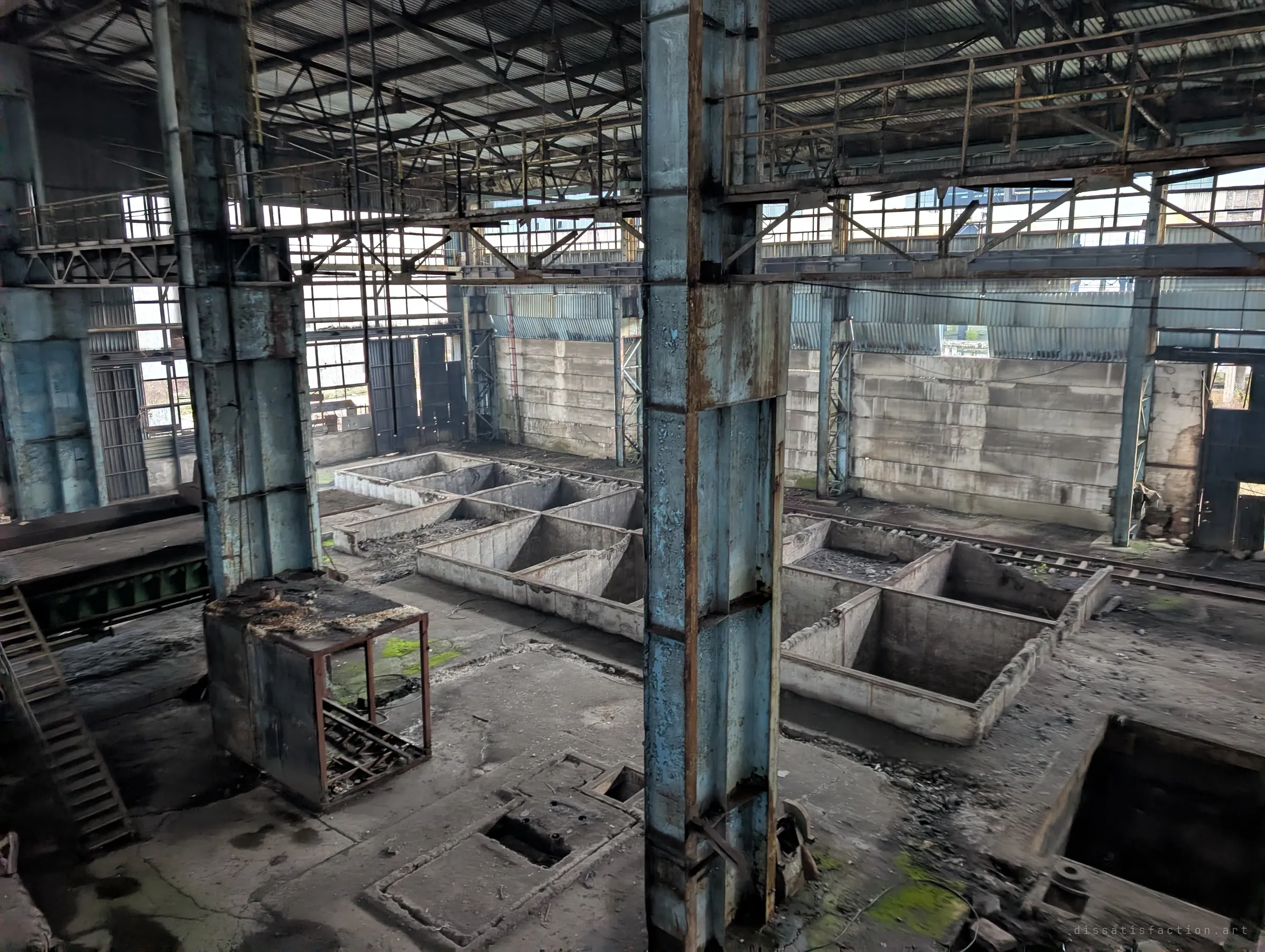 Looking down from a raised level onto rows of empty concrete bunkers and rail tracks between peeling blue steel columns under the roof trusses.