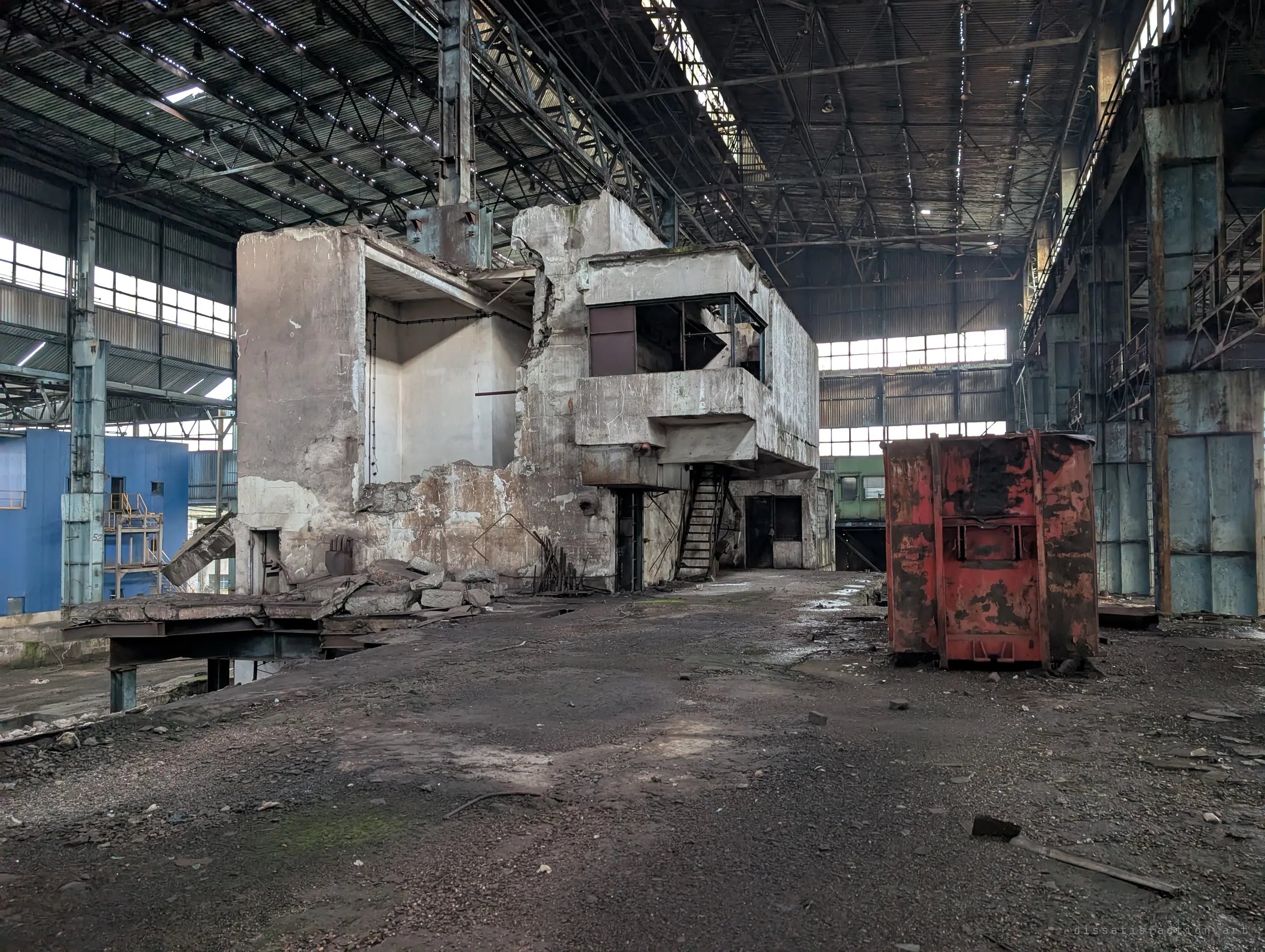 A half-demolished concrete control structure stands in the middle of the hall beside a rust-red skip, dwarfed by the steel roof and window bands.