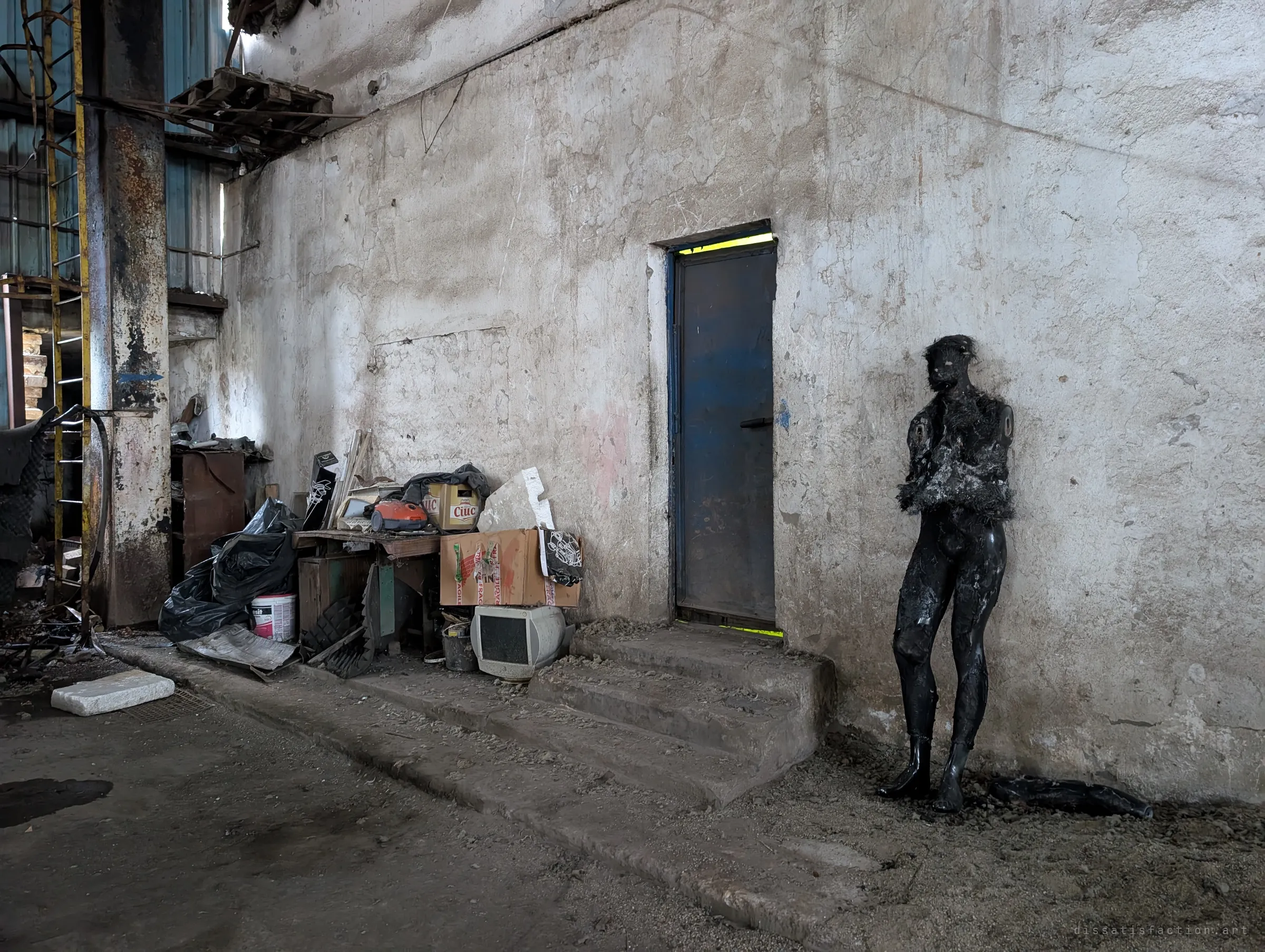 A blackened, fur-draped mannequin leans against a pale wall beside a blue steel door, next to a pile of recent junk: boxes, a vacuum cleaner and an old CRT monitor.