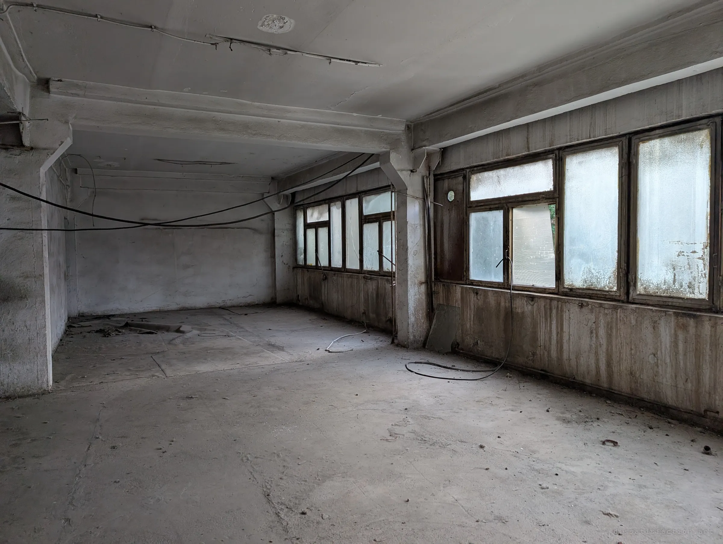 An empty office floor with bare concrete, sagging cables and a long run of frosted windows along one wall.