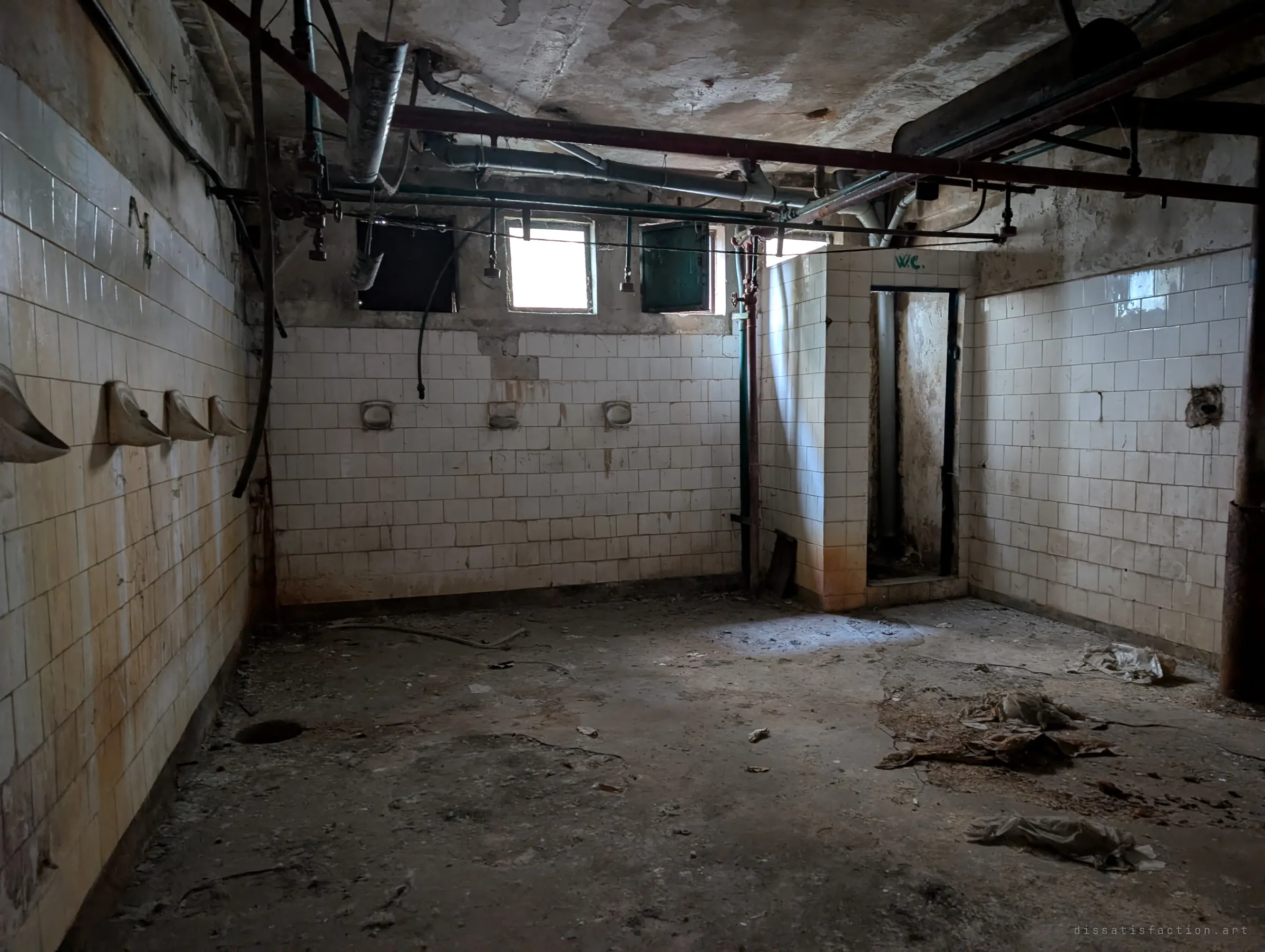A tiled workers' washroom with a row of wall urinals, stripped shower pipes overhead and a small toilet cubicle, lit by high windows.