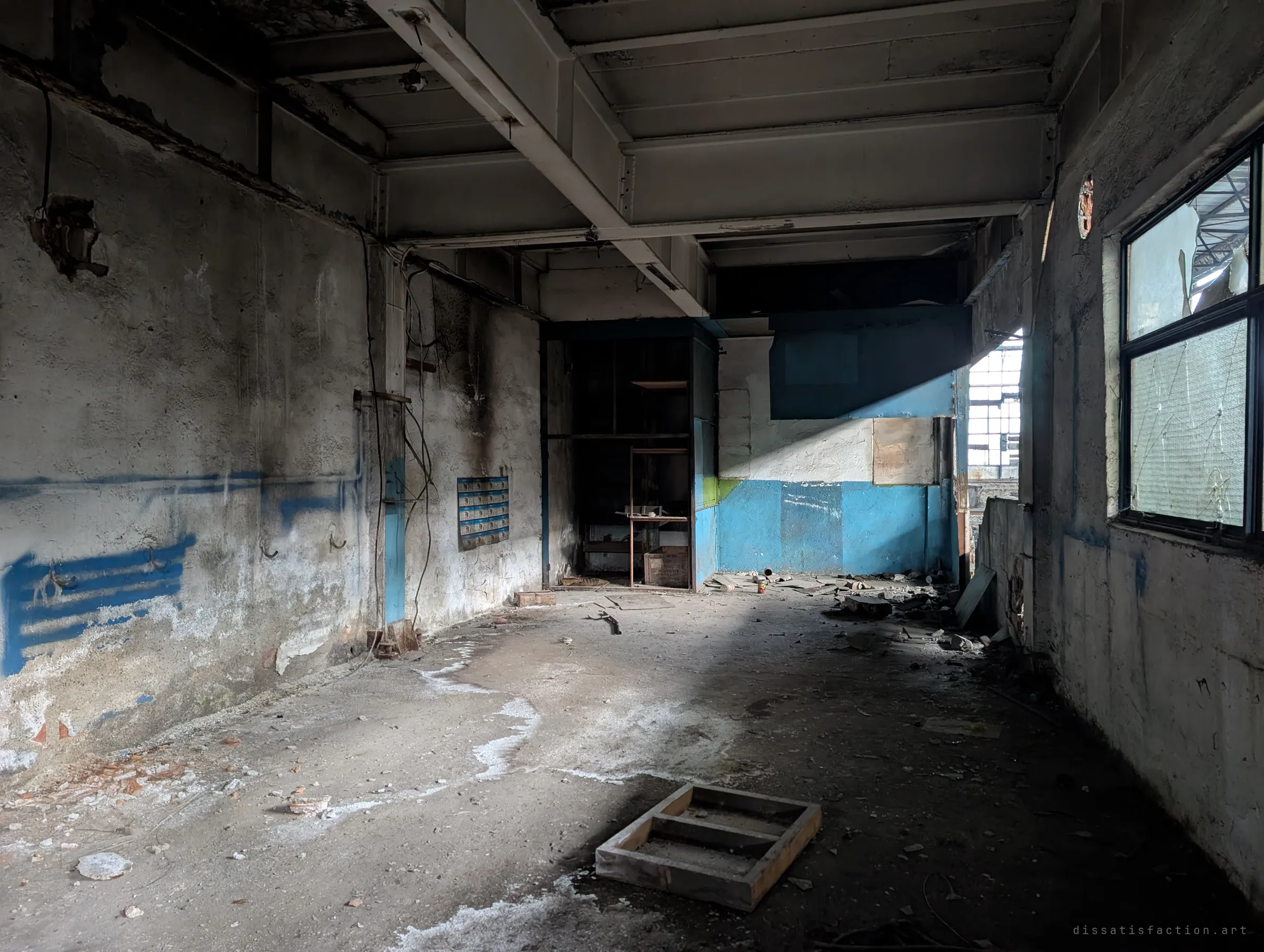 A soot-streaked upper-floor room with blue-painted walls, a steel-beam ceiling and a light shaft cutting across the debris-strewn floor. A broken wired-glass window on the right looks back into the main hall.