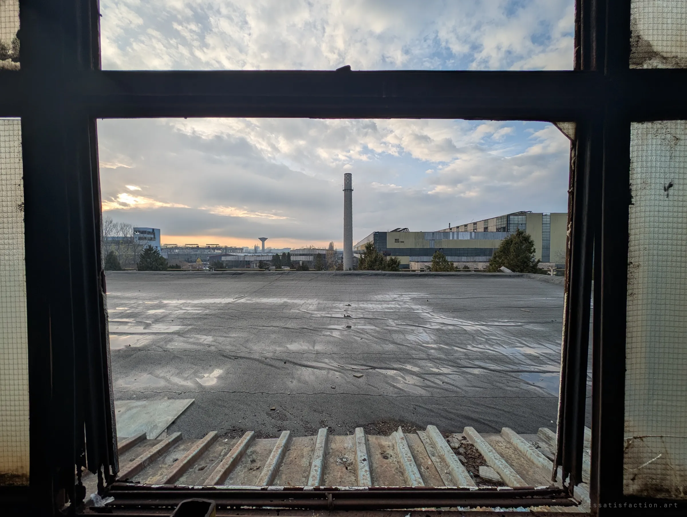 Looking out through a rusted, wire-glass window frame onto a wet flat roof, with a lone chimney and neighbouring factory halls under a dusk sky. The window frame turns the empty roof into a stage.